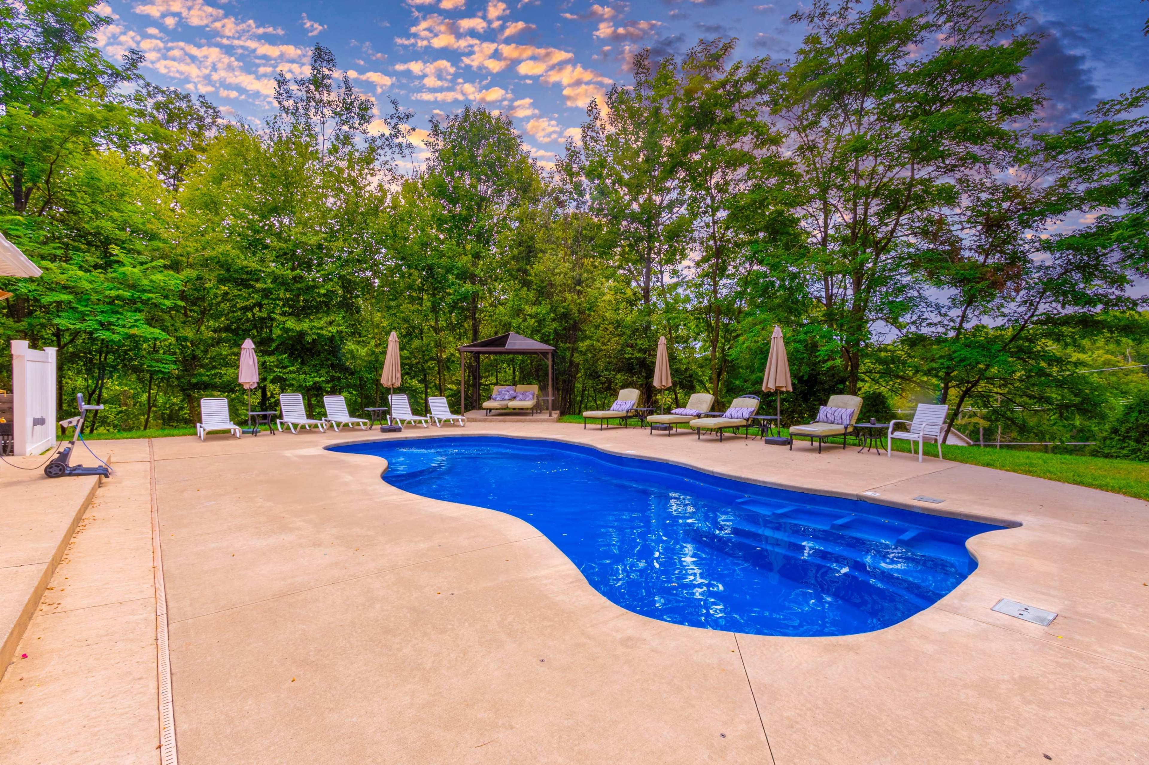 The image shows a residential pool surrounded by lounge chairs and umbrellas, set against a backdrop of trees and a colorful sky.