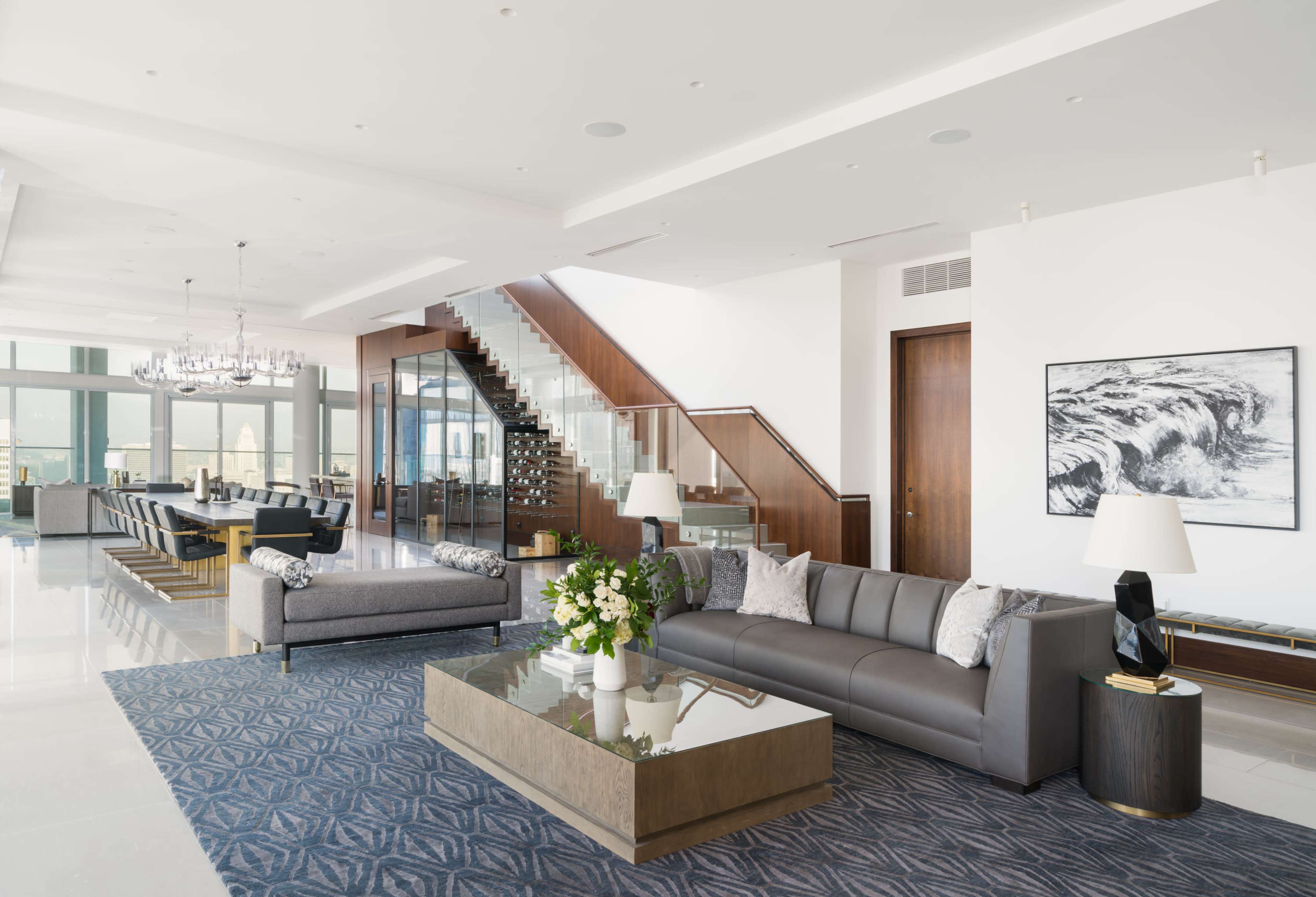 The image shows a modern, spacious living room with a two-story ceiling, featuring a gray sofa, a glass coffee table, and a staircase that leads to an upper level.
