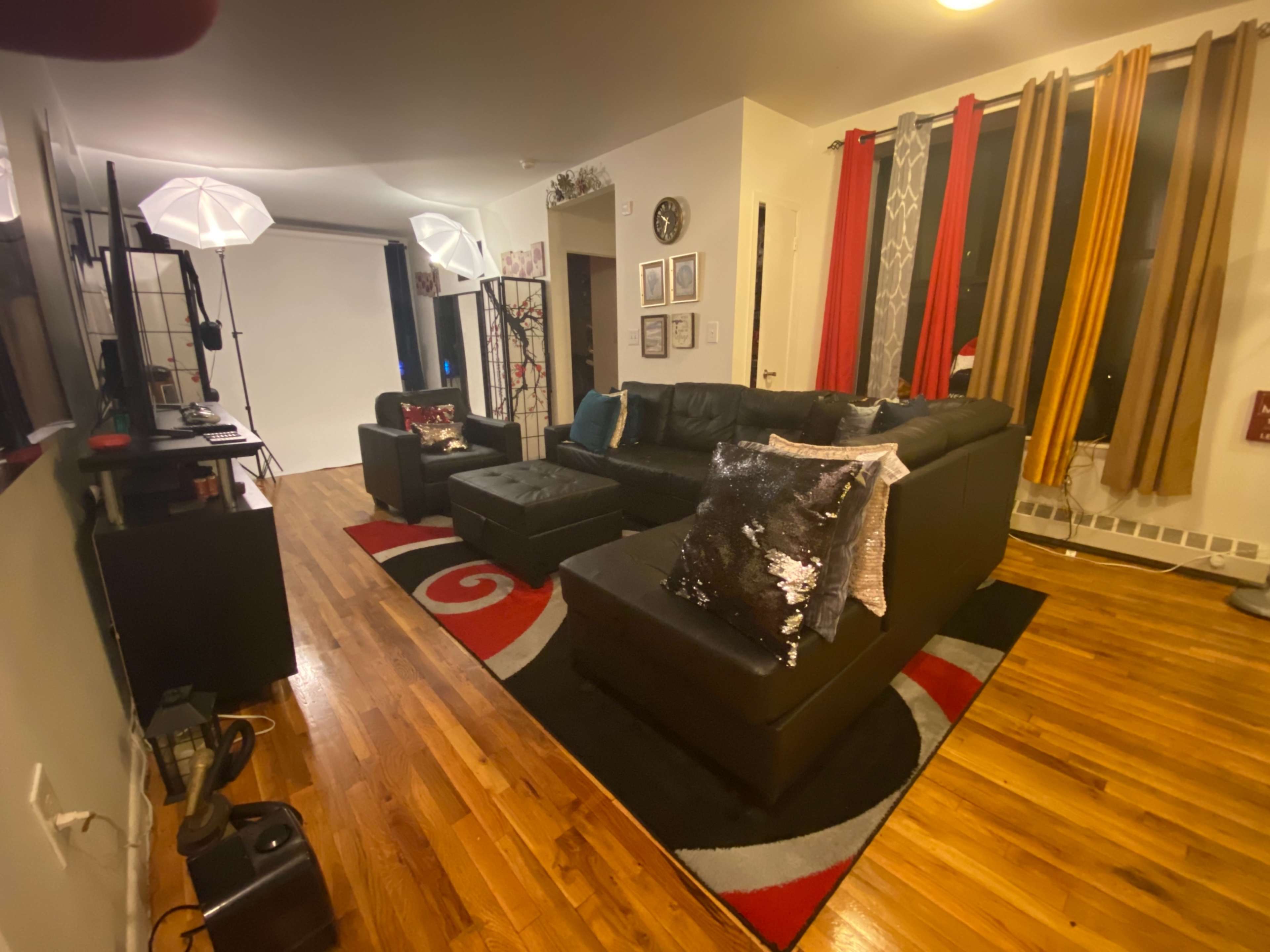 The image shows a living room with a black sectional sofa, colorful curtains, and a modern decorative rug on a wooden floor.