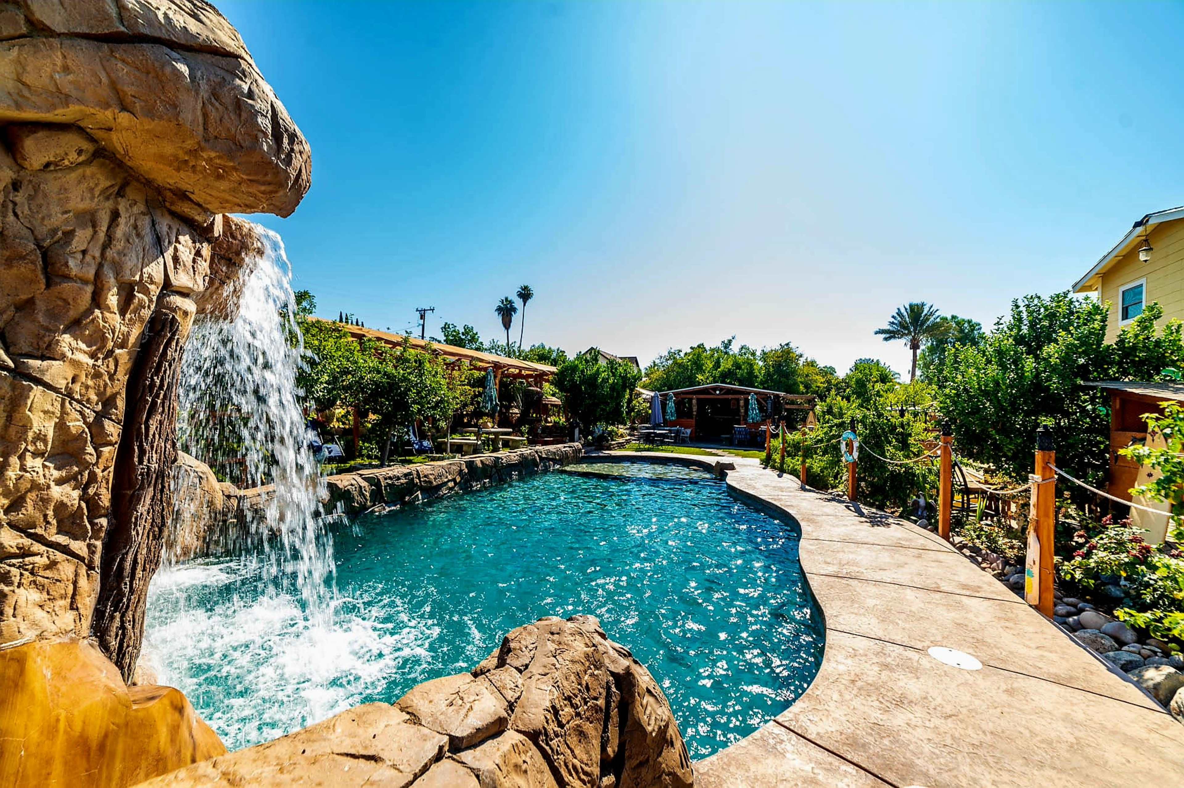 The image displays a pool surrounded by rock formations, a waterfall, and lush greenery under a clear blue sky.