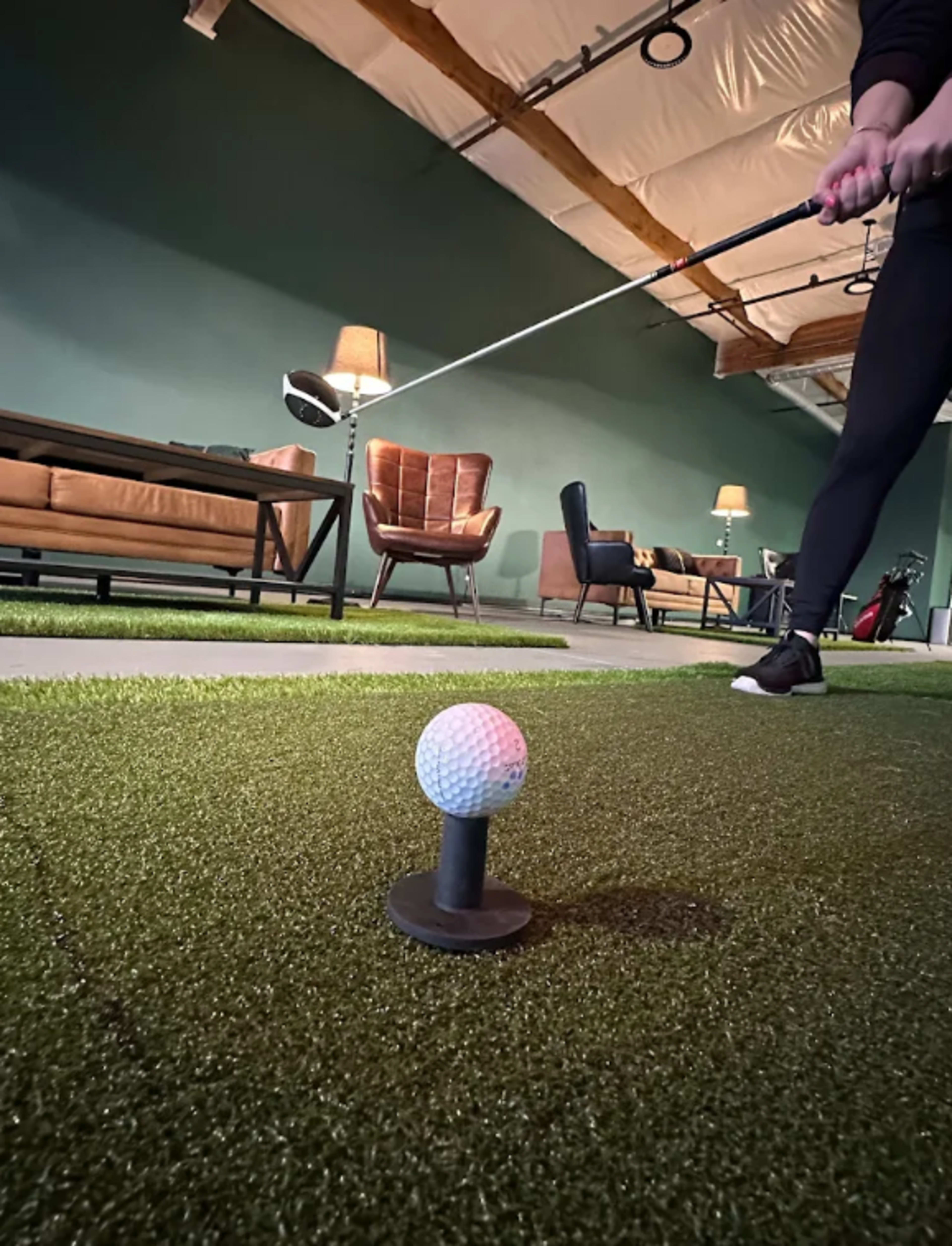 A golfer prepares to hit a golf ball teed up on a green artificial surface inside a room with furniture and lamps in the background.