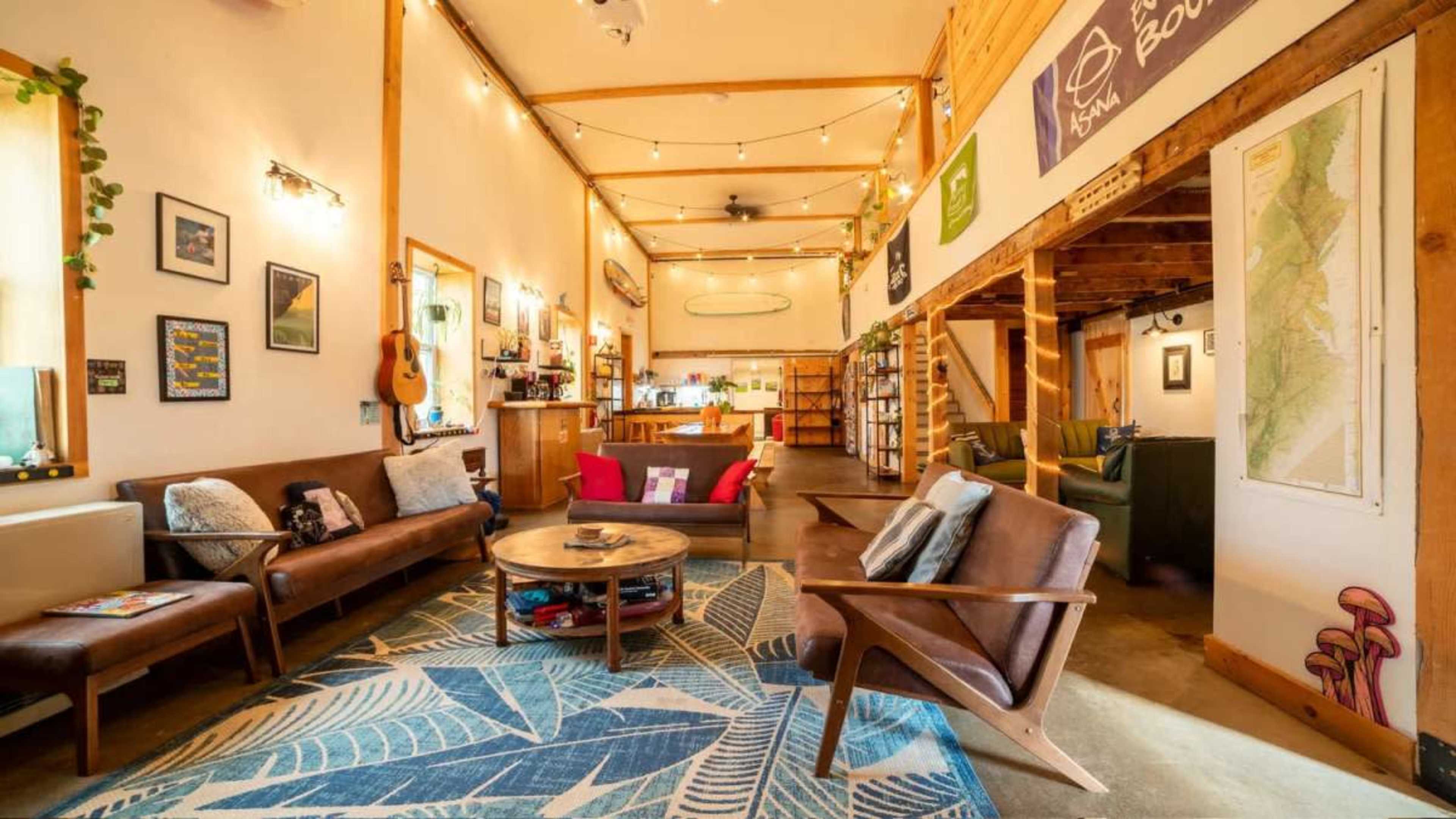 The image shows a cozy, well-lit common area with wooden walls, comfortable seating, a round coffee table, and a vibrant area rug, featuring shelves filled with books and various decorations.