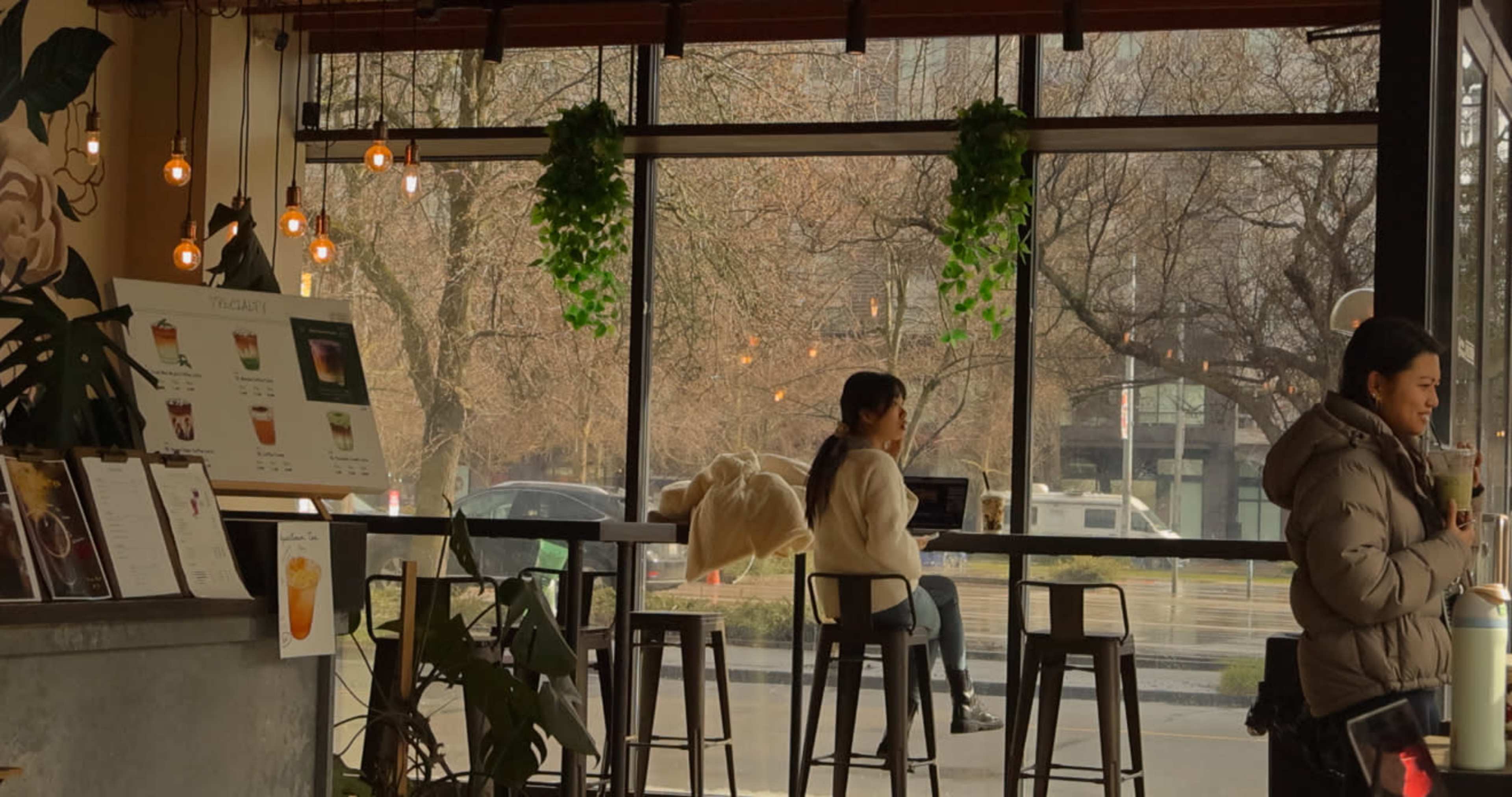 A café interior features a glass wall with outdoor views, showcasing customers seated at high stools and others standing, surrounded by hanging plants and warm lighting.