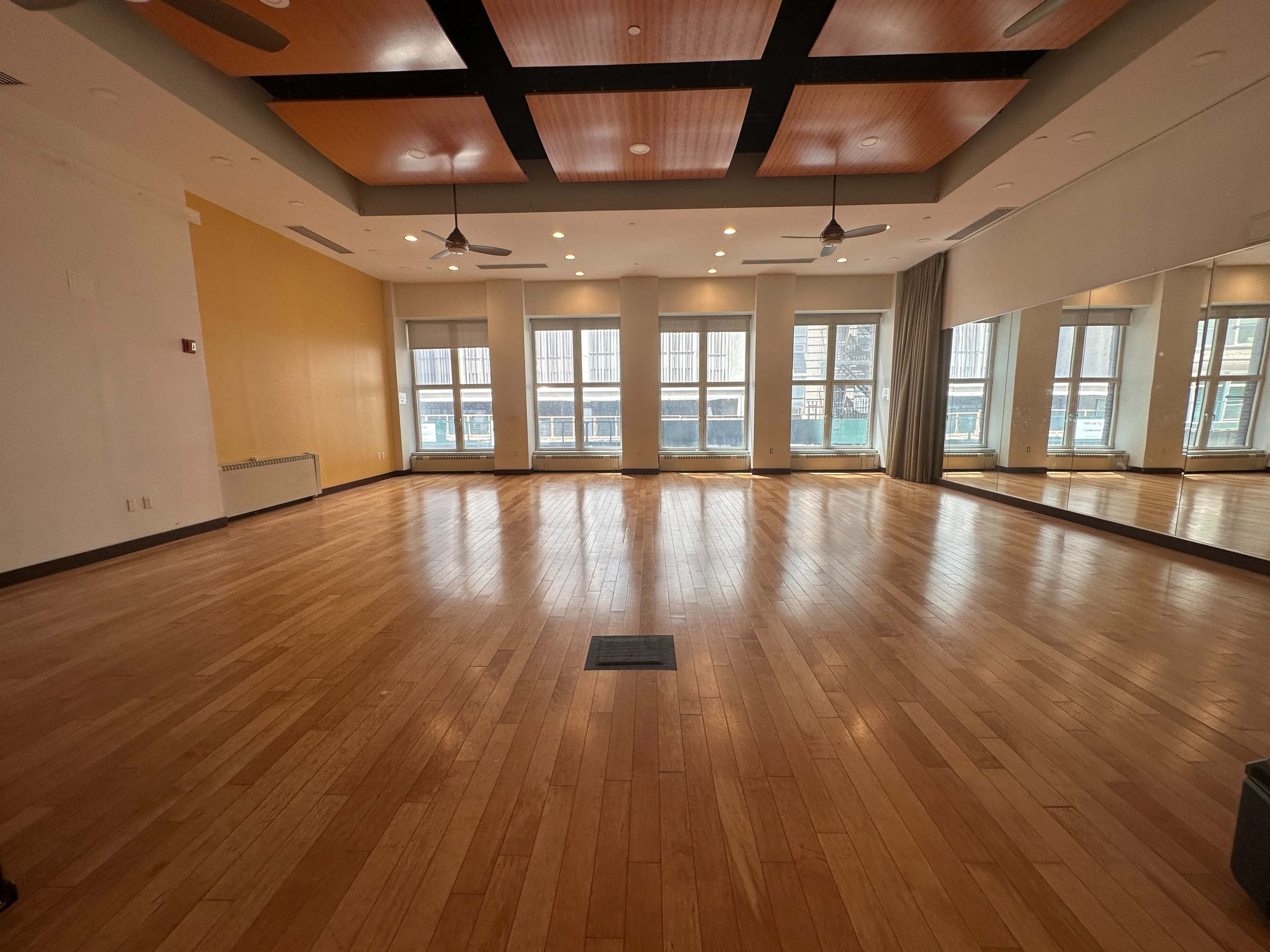 An empty wooden-floored room with large windows and a mirrored wall, featuring ceiling fans and a paneled ceiling design.