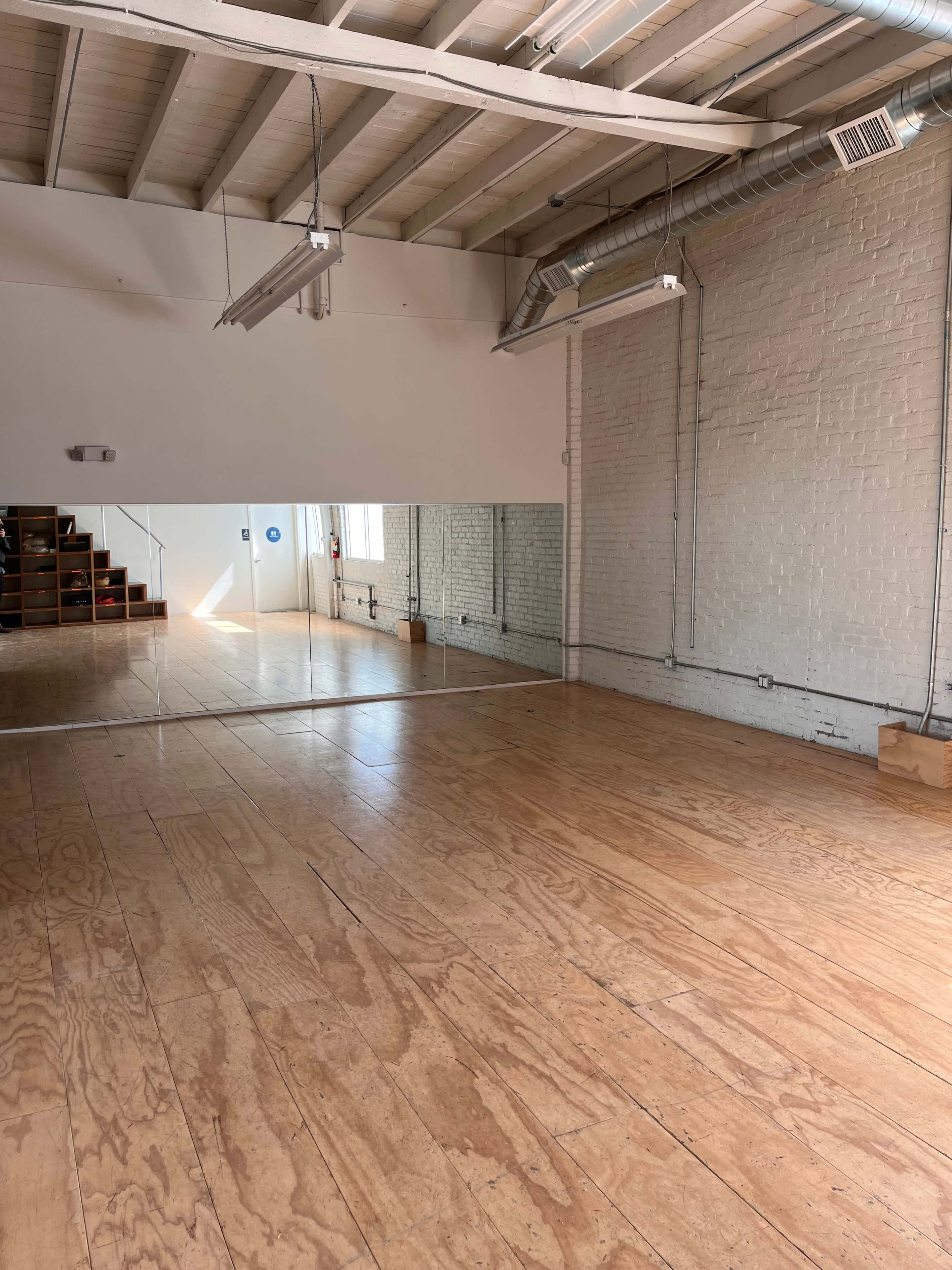 A spacious room with wooden floors and a large mirror along one wall, featuring exposed brick and pipes.