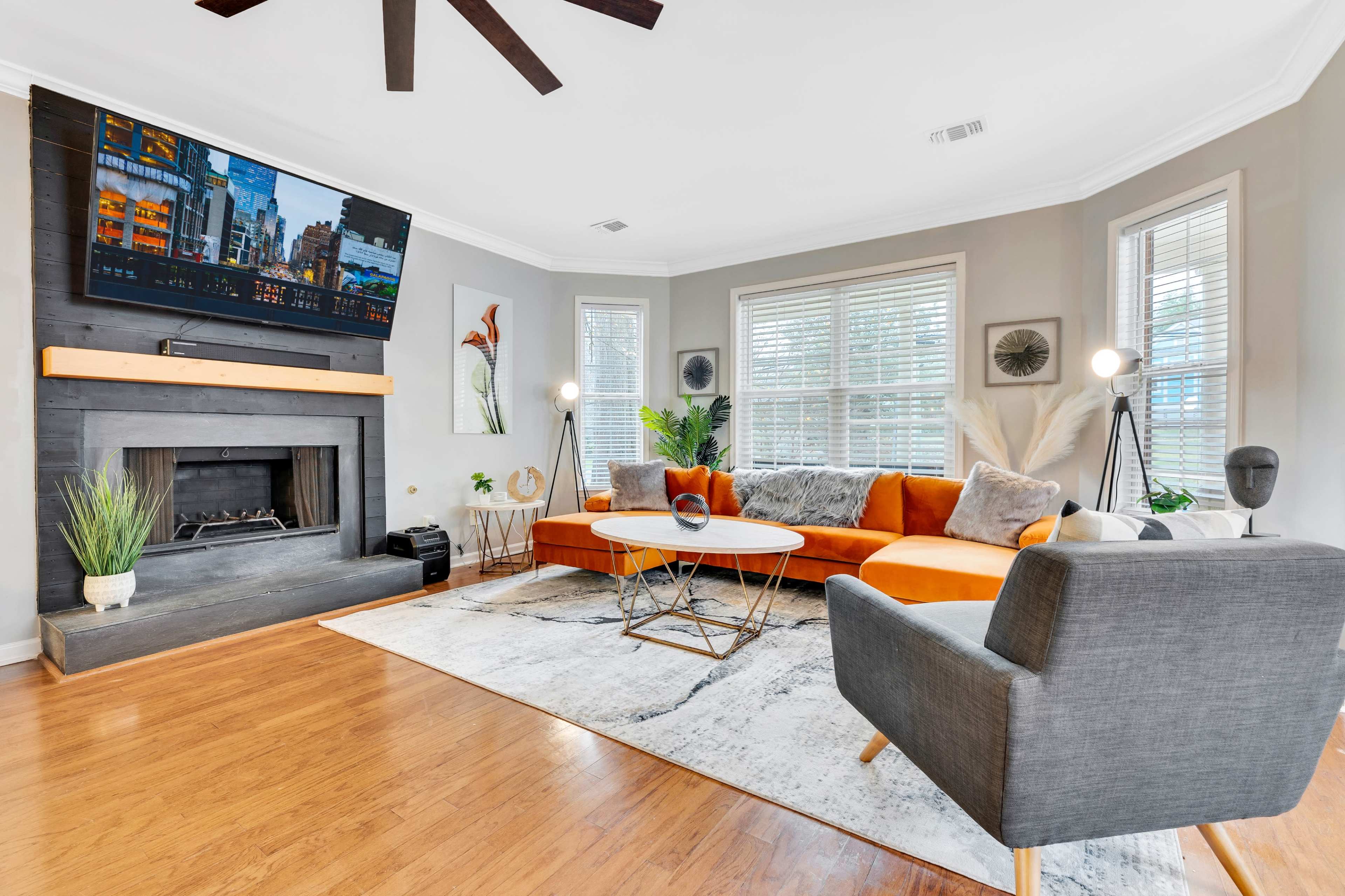 The image shows a living room with an orange sectional sofa, a round coffee table, a modern chair, and a flat-screen TV mounted above a fireplace.