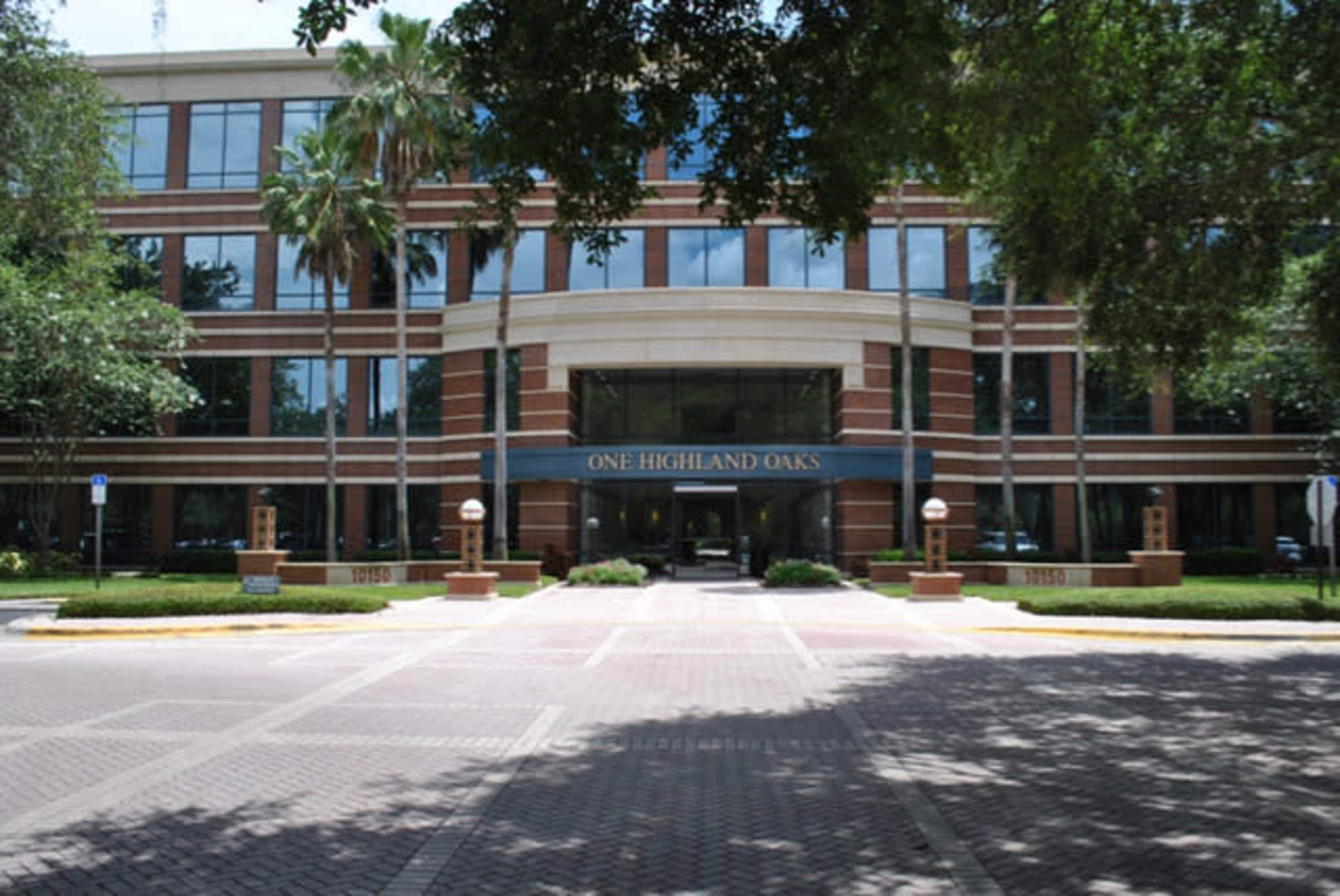 A modern office building with a prominent sign reading "One Highland Oaks" surrounded by palm trees and a paved entrance.