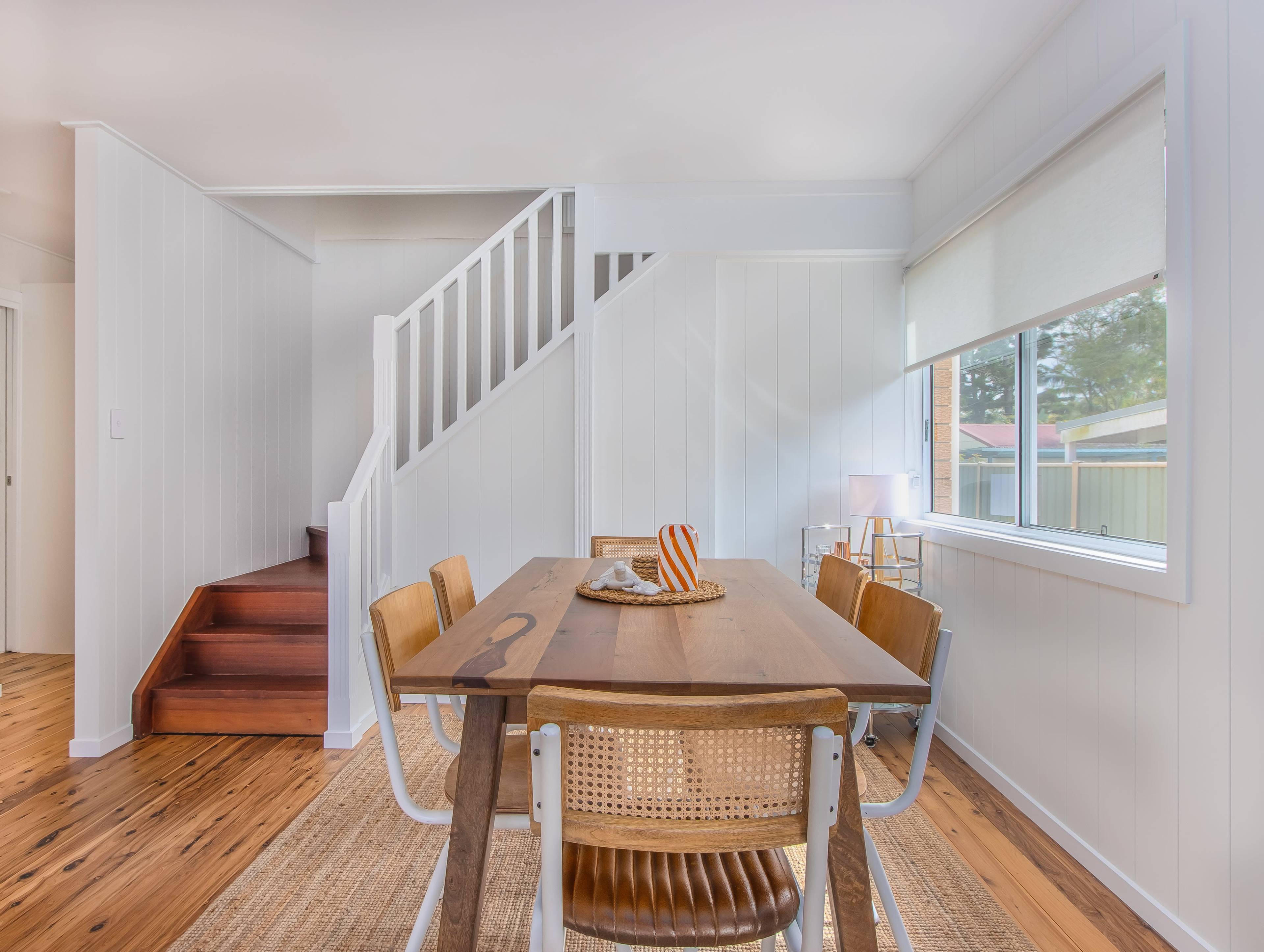 A dining area features a wooden table surrounded by chairs, with a staircase and window in the background.