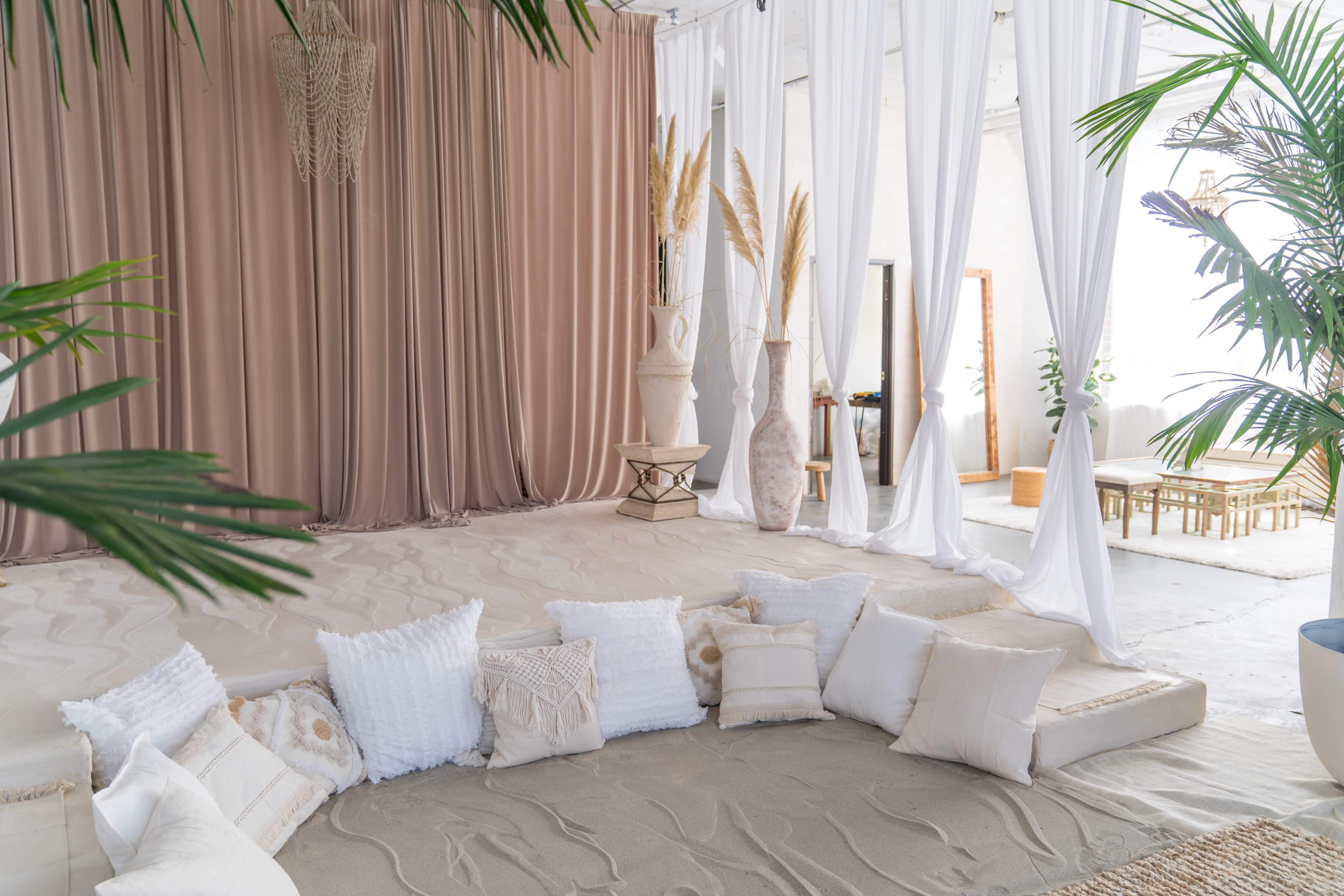 The image shows a cozy indoor space with a circular seating area made of cushions, surrounded by draped fabric, soft flooring, and decorative plants.
