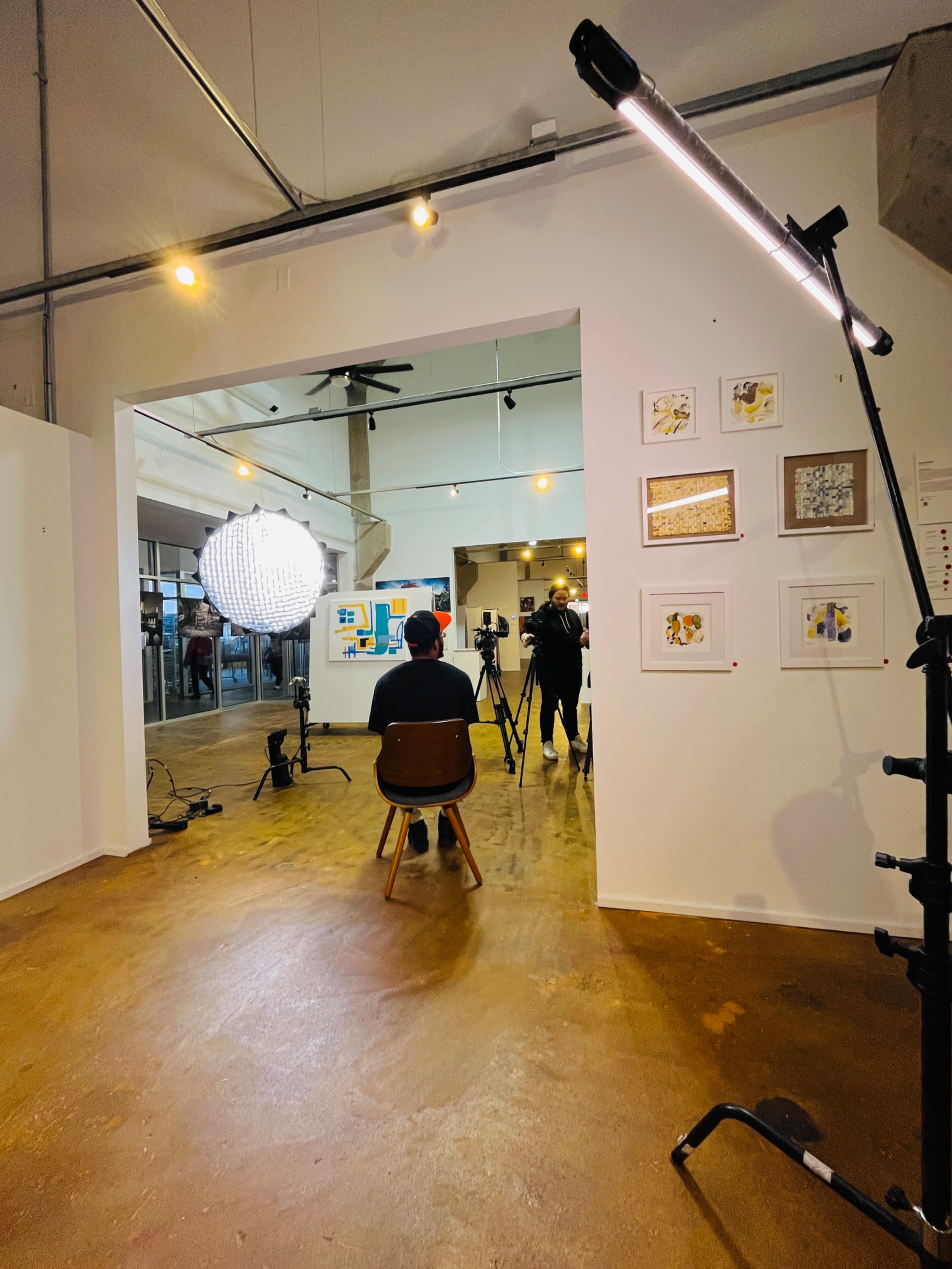 A person sits on a chair in an art gallery, while two cameras and a large light are positioned nearby.