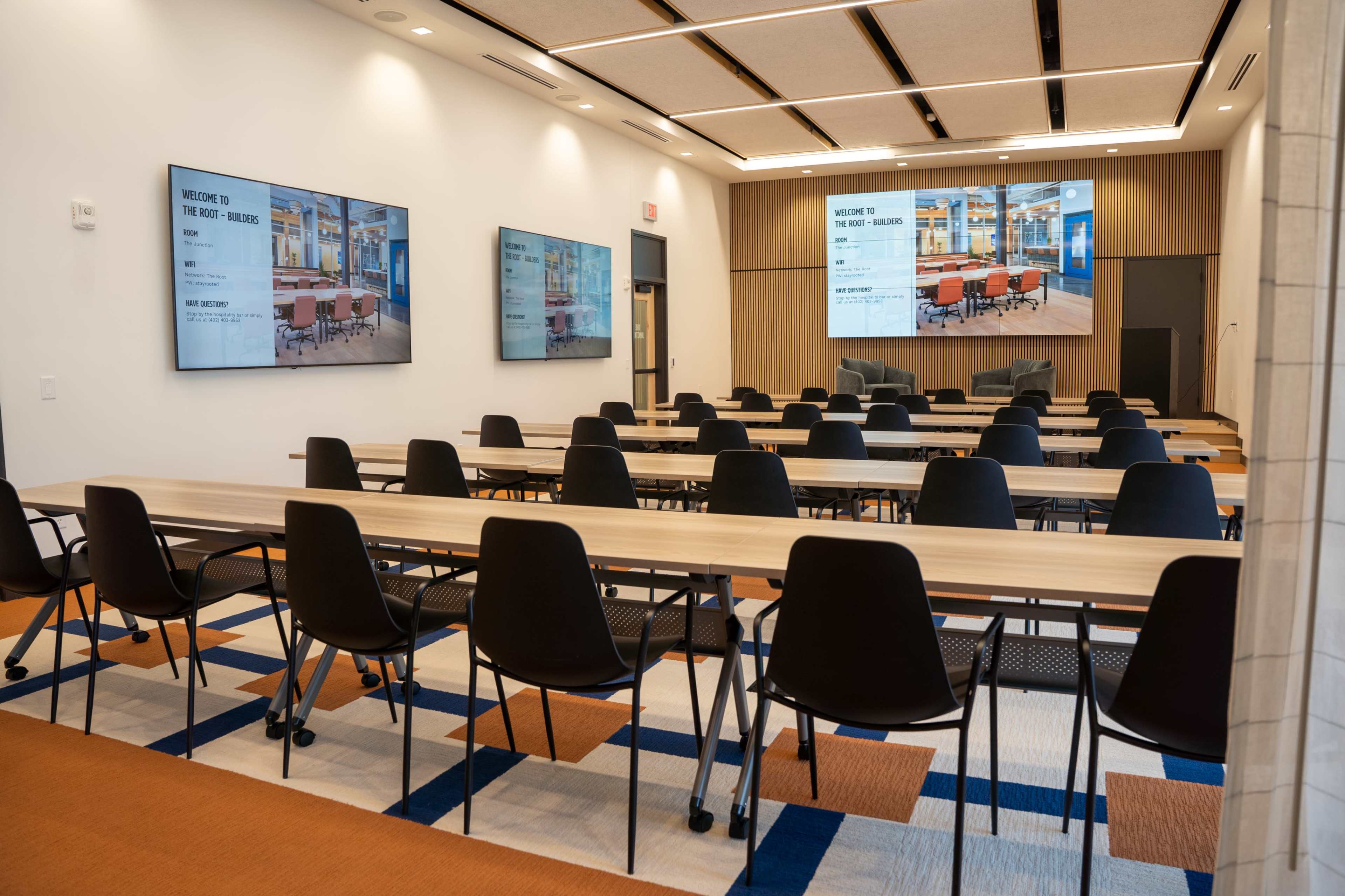 The image shows a modern conference room with long tables arranged in rows, black chairs, and two large screens displaying welcome messages on the wall.