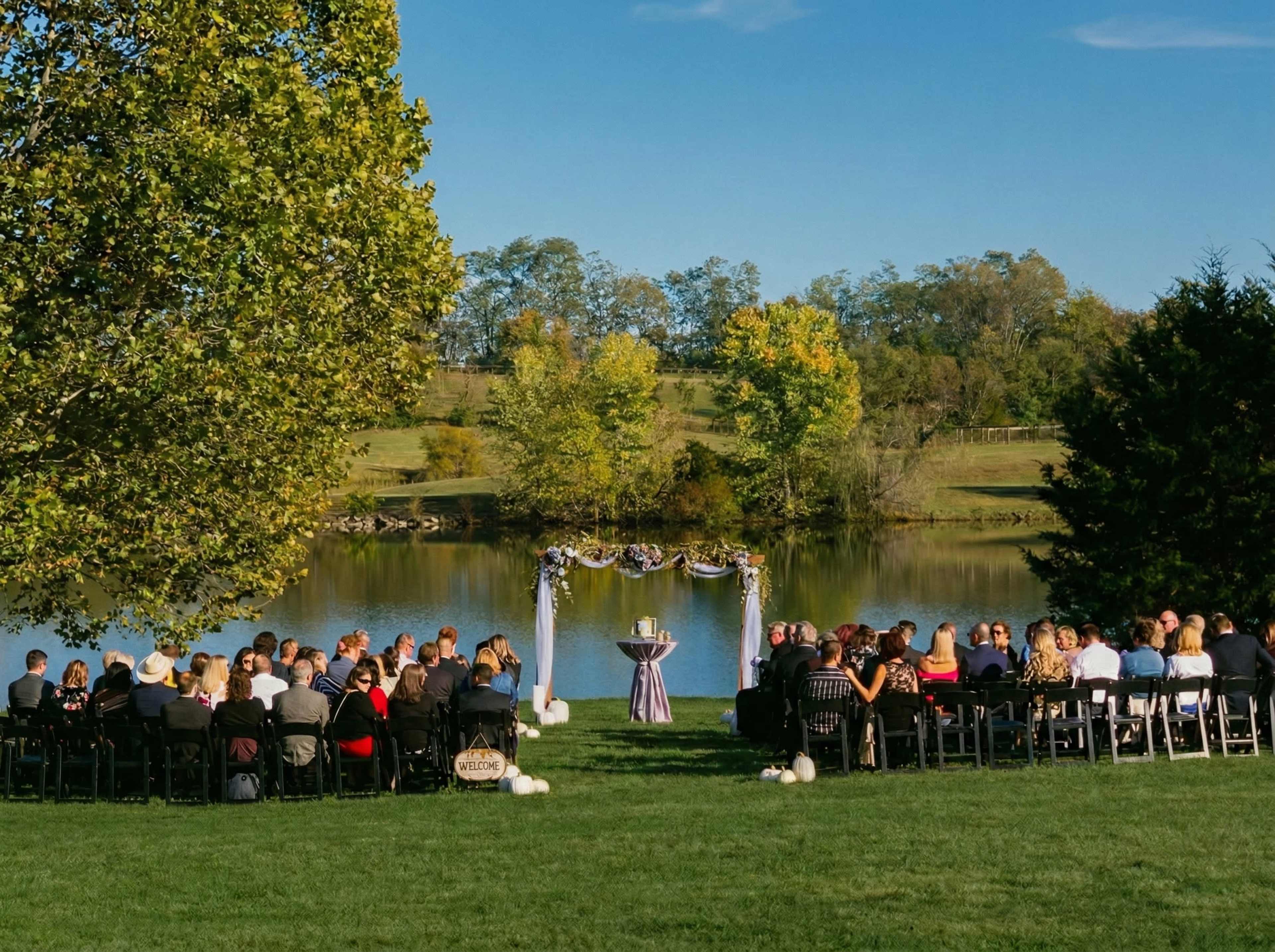 A wedding ceremony is set up by a lake, with guests seated in black chairs and a floral arch at the front.