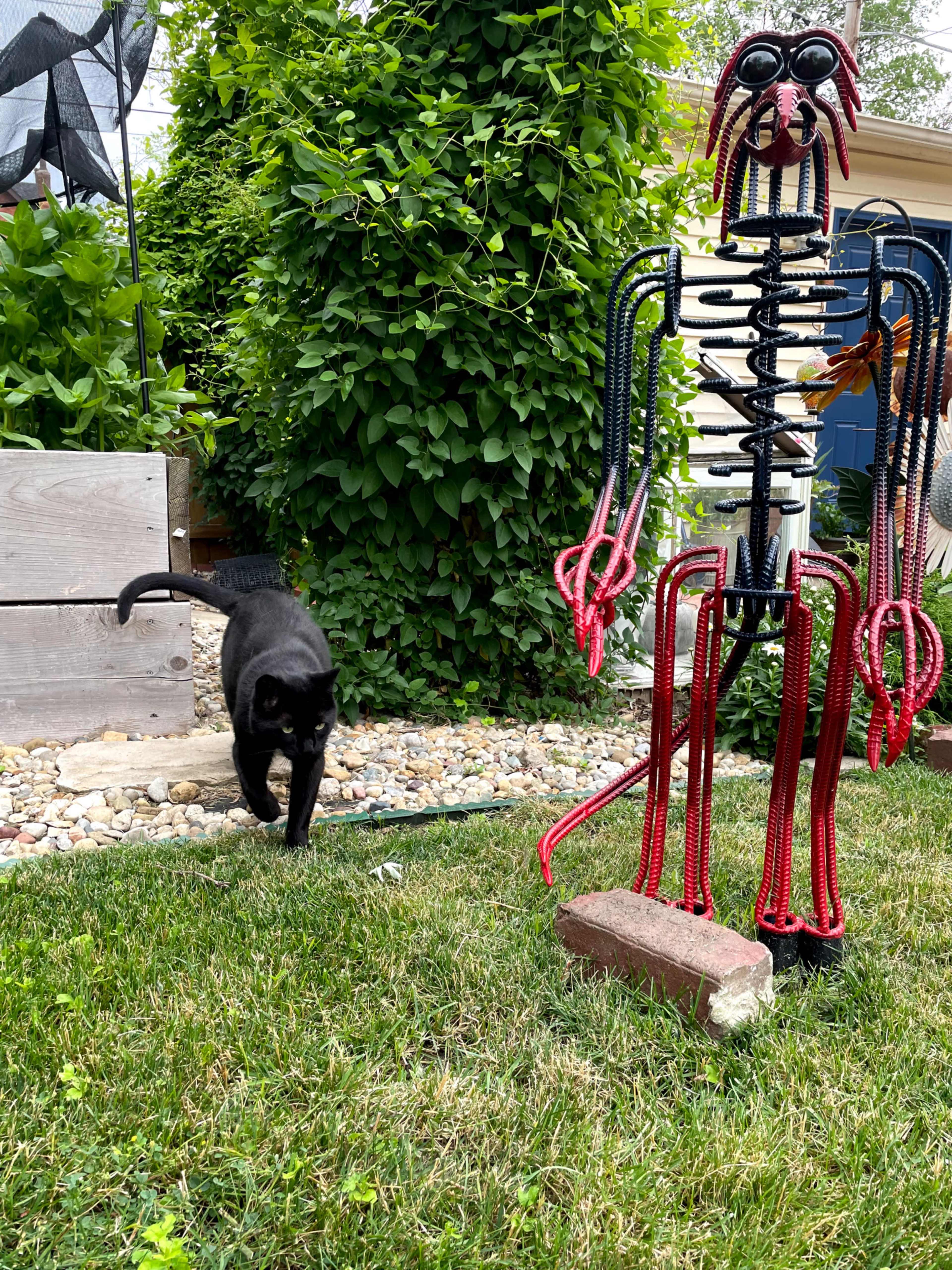 A black cat walks past a colorful metal sculpture of a figure in a grassy garden.