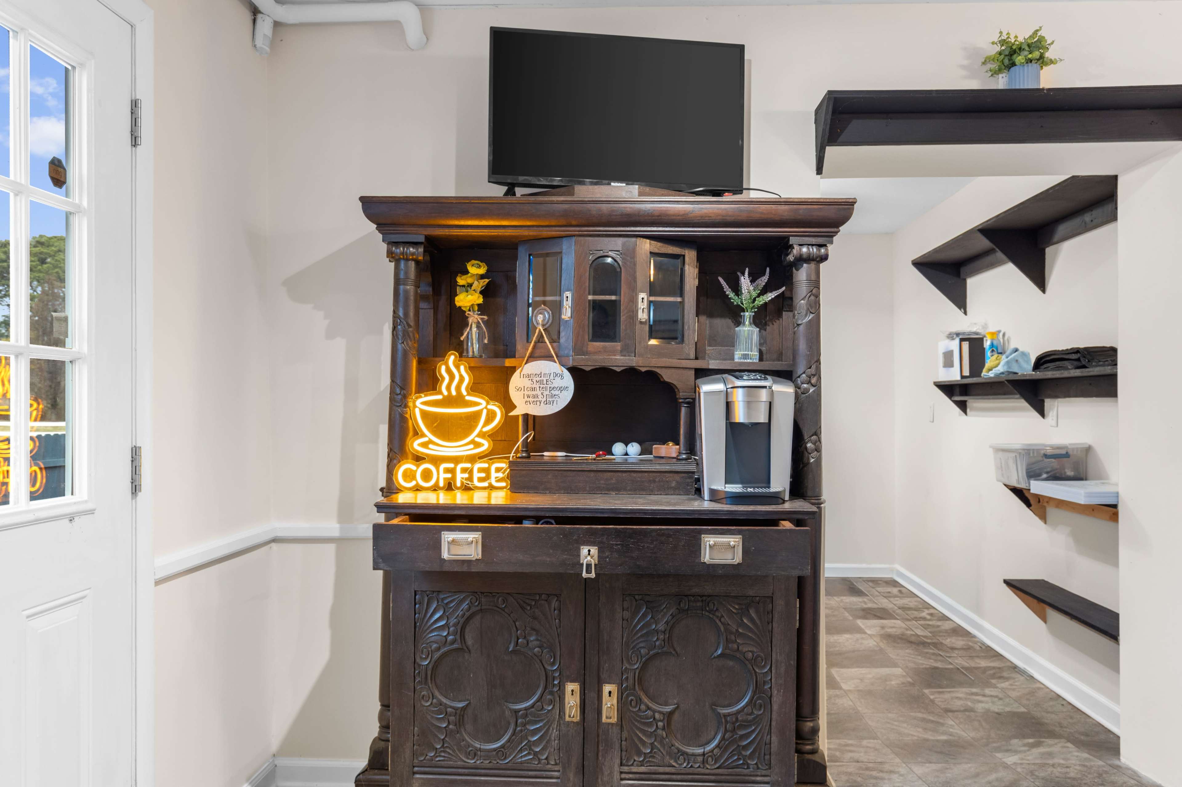 A wooden cabinet with a coffee maker and a neon coffee sign is positioned against a wall, complemented by a television mounted above it.