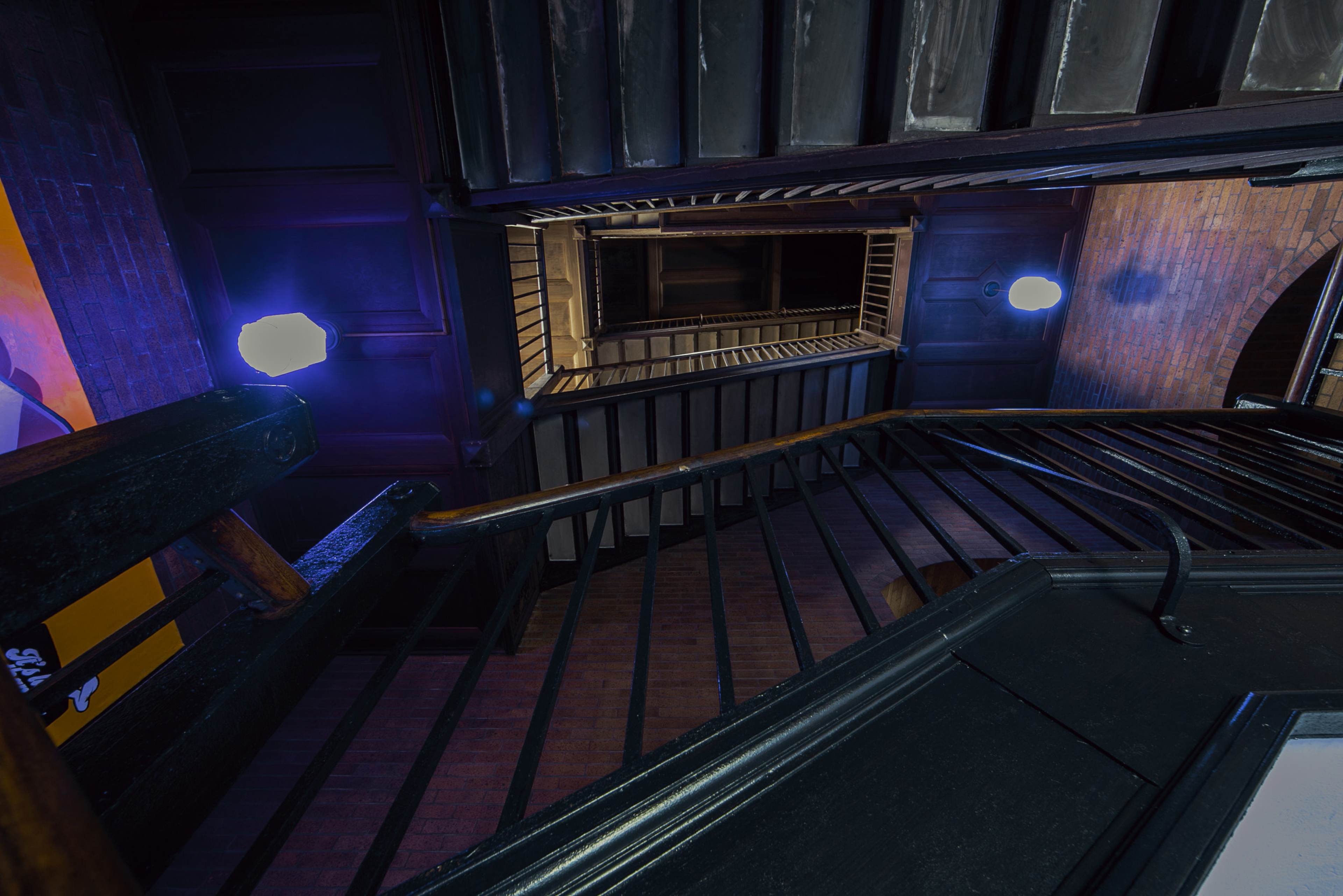 The image shows a downward view of a staircase with wooden railings, illuminated by wall-mounted lights.