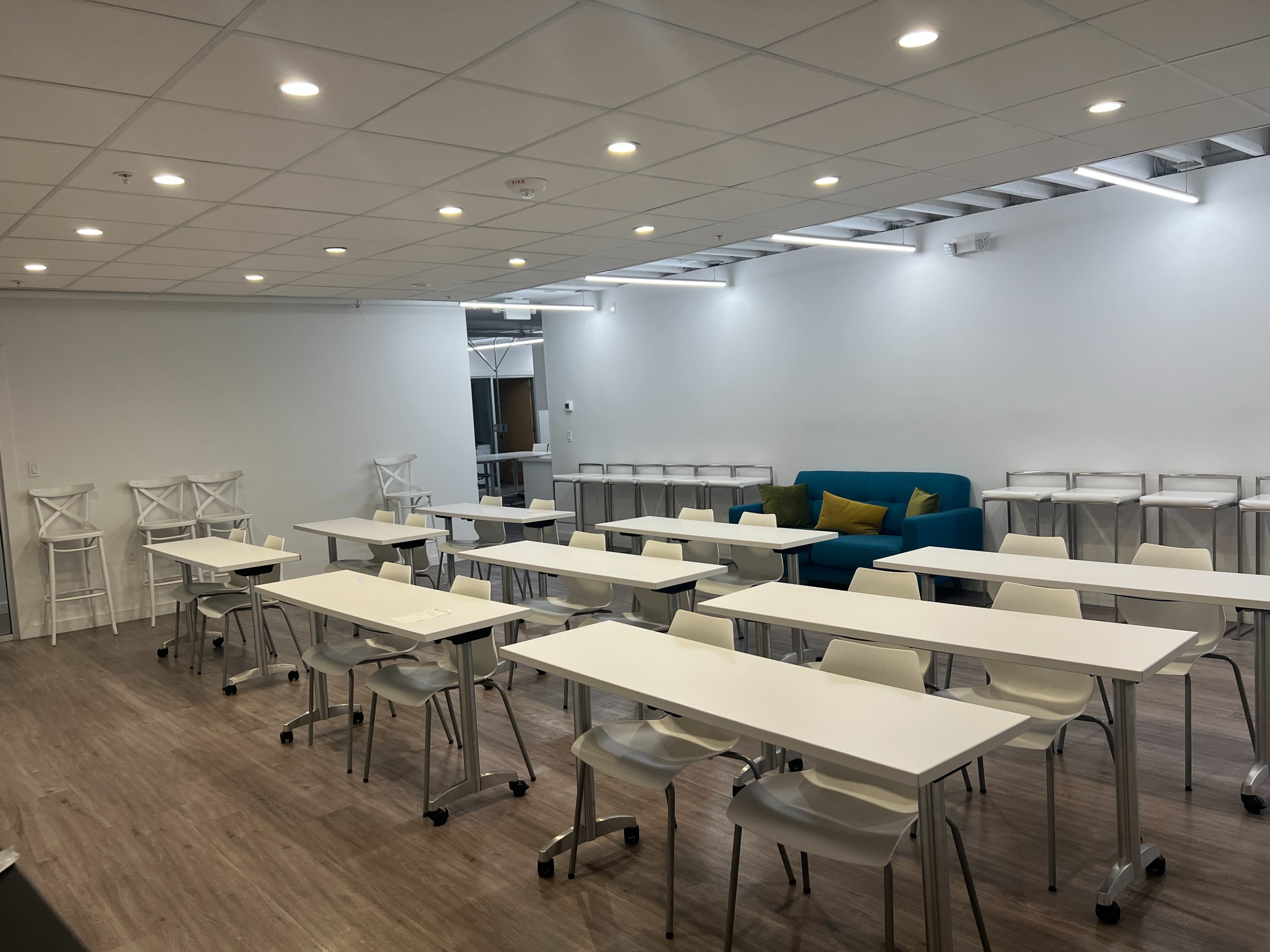 A modern classroom setup with rows of white tables and chairs, a blue couch in the corner, and bright, well-lit walls.