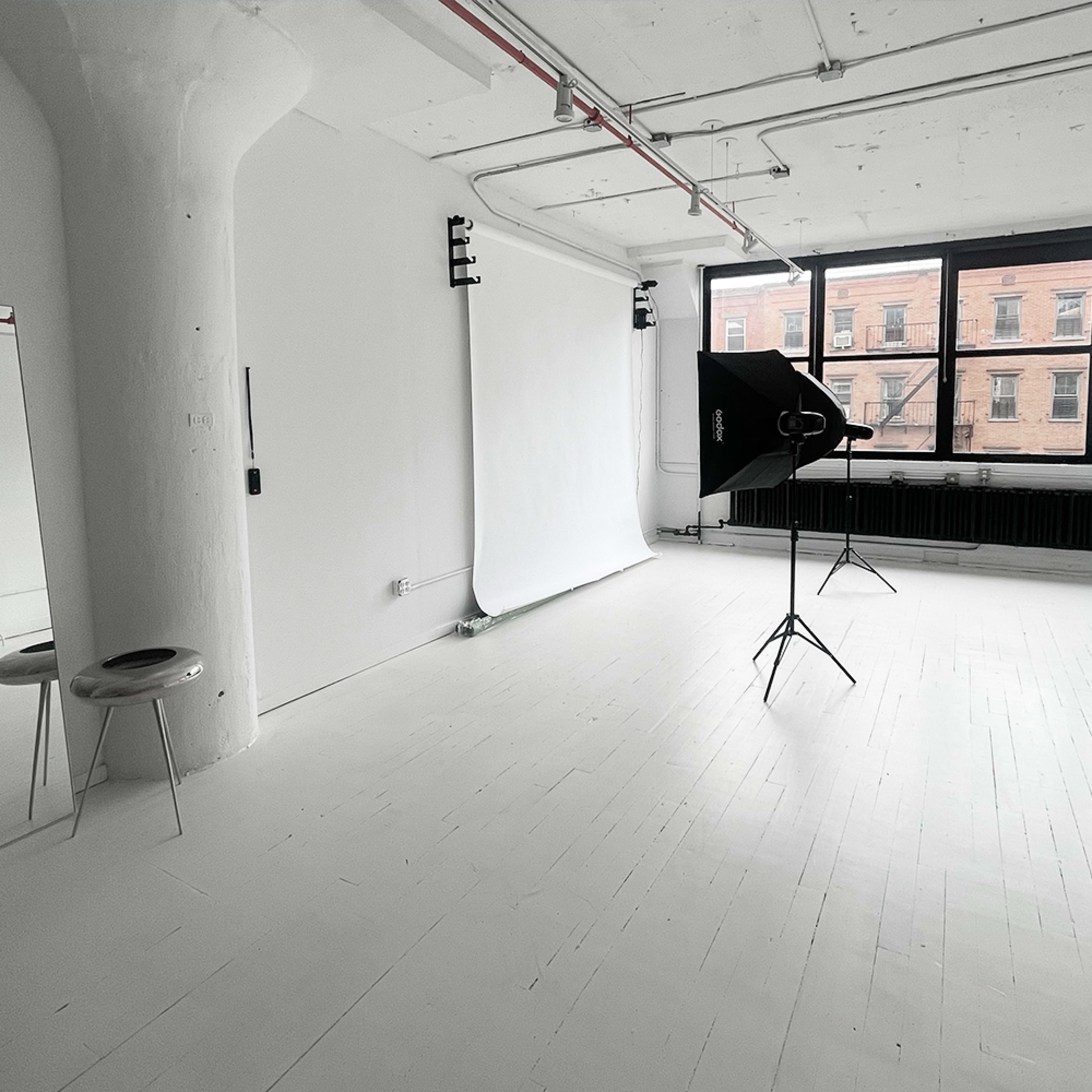 A minimalist photo studio with a white backdrop, lighting equipment, and large windows letting in natural light.