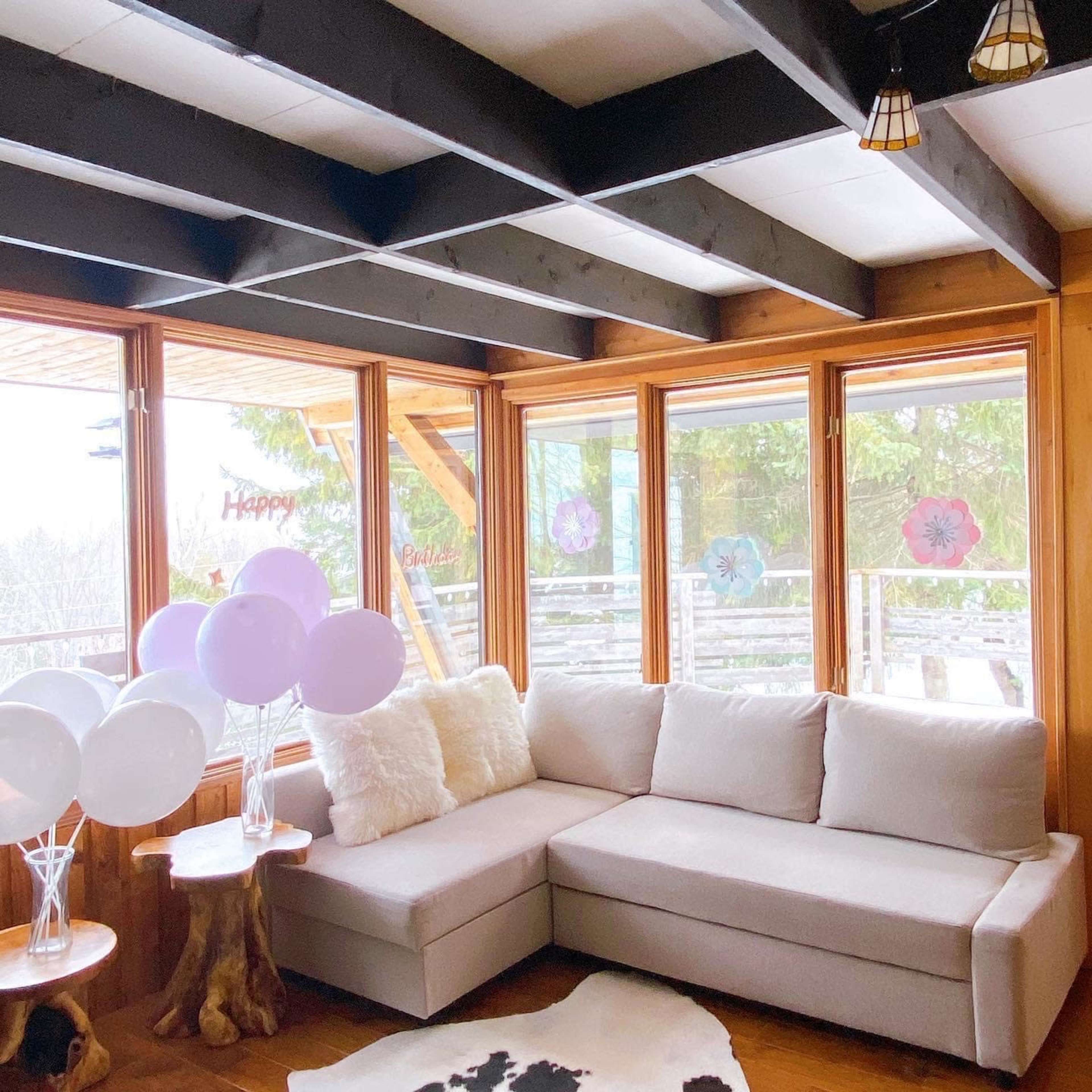A bright sunroom features a beige sectional sofa, decorative balloons, and wooden accents with large windows overlooking a greenery-filled view.