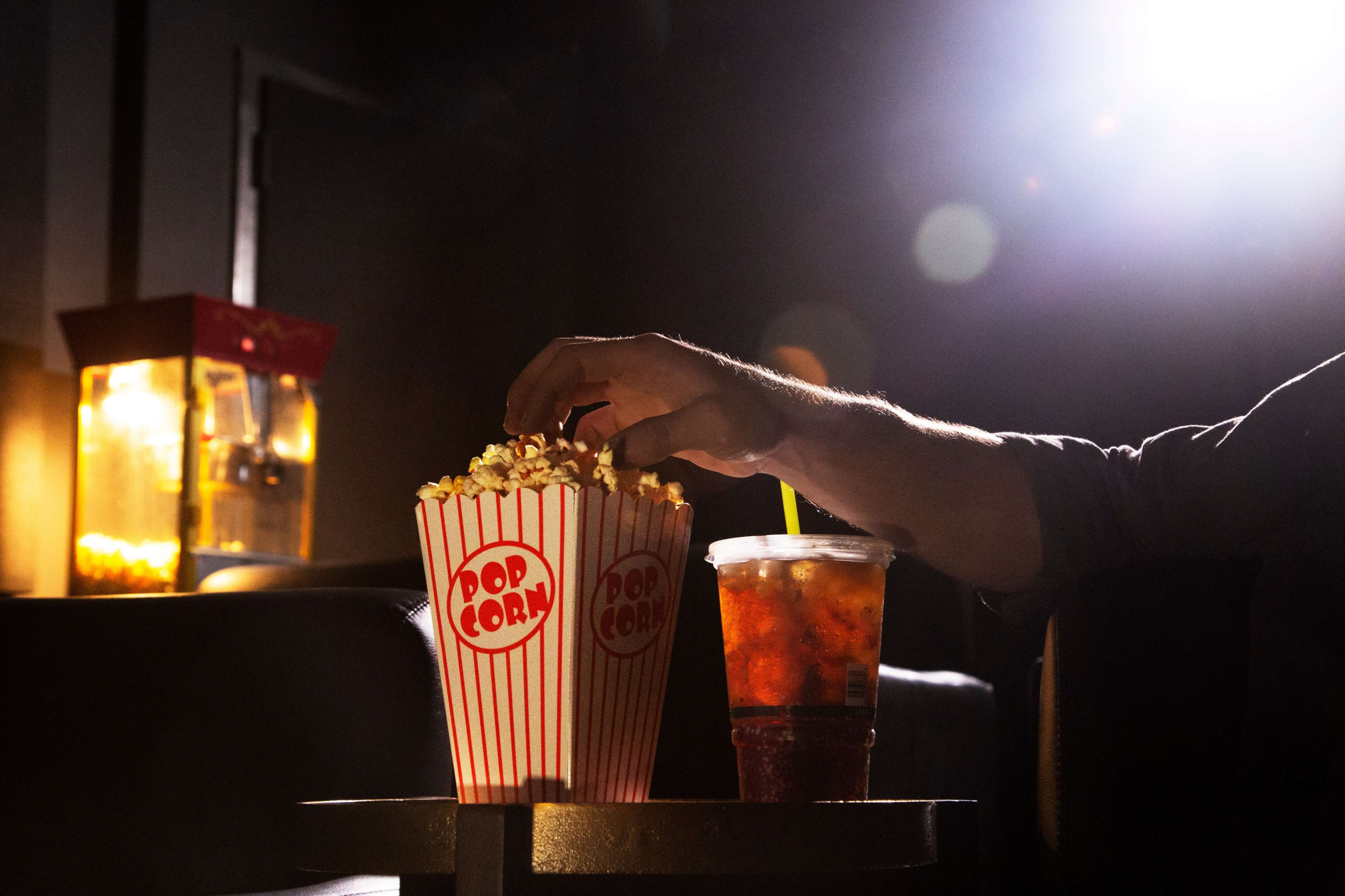 A hand reaches into a large striped popcorn bucket next to a cup of iced drink, set against a dimly lit background.