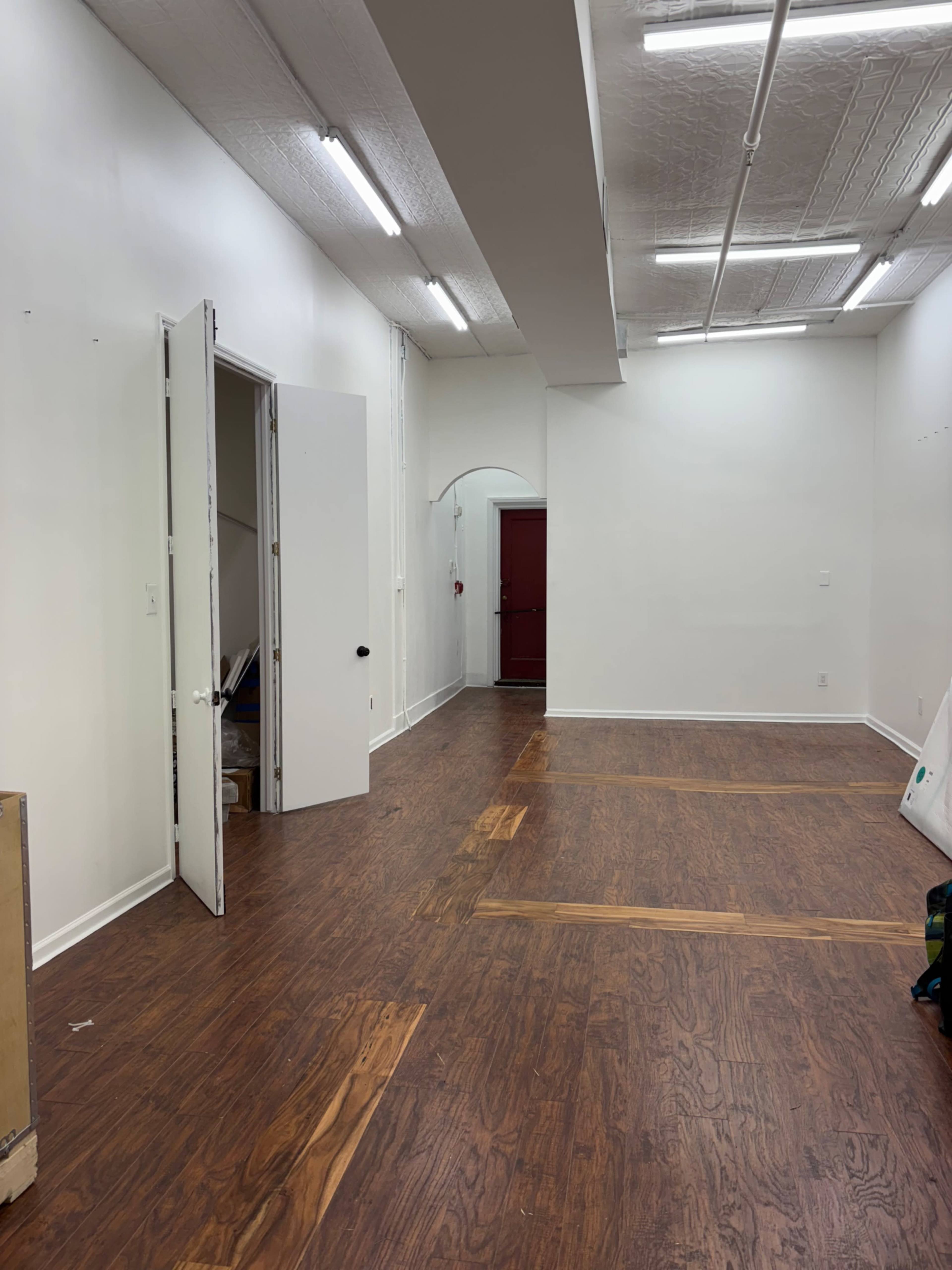 The image shows an empty room with wooden flooring, white walls, and an open door leading to a hallway.