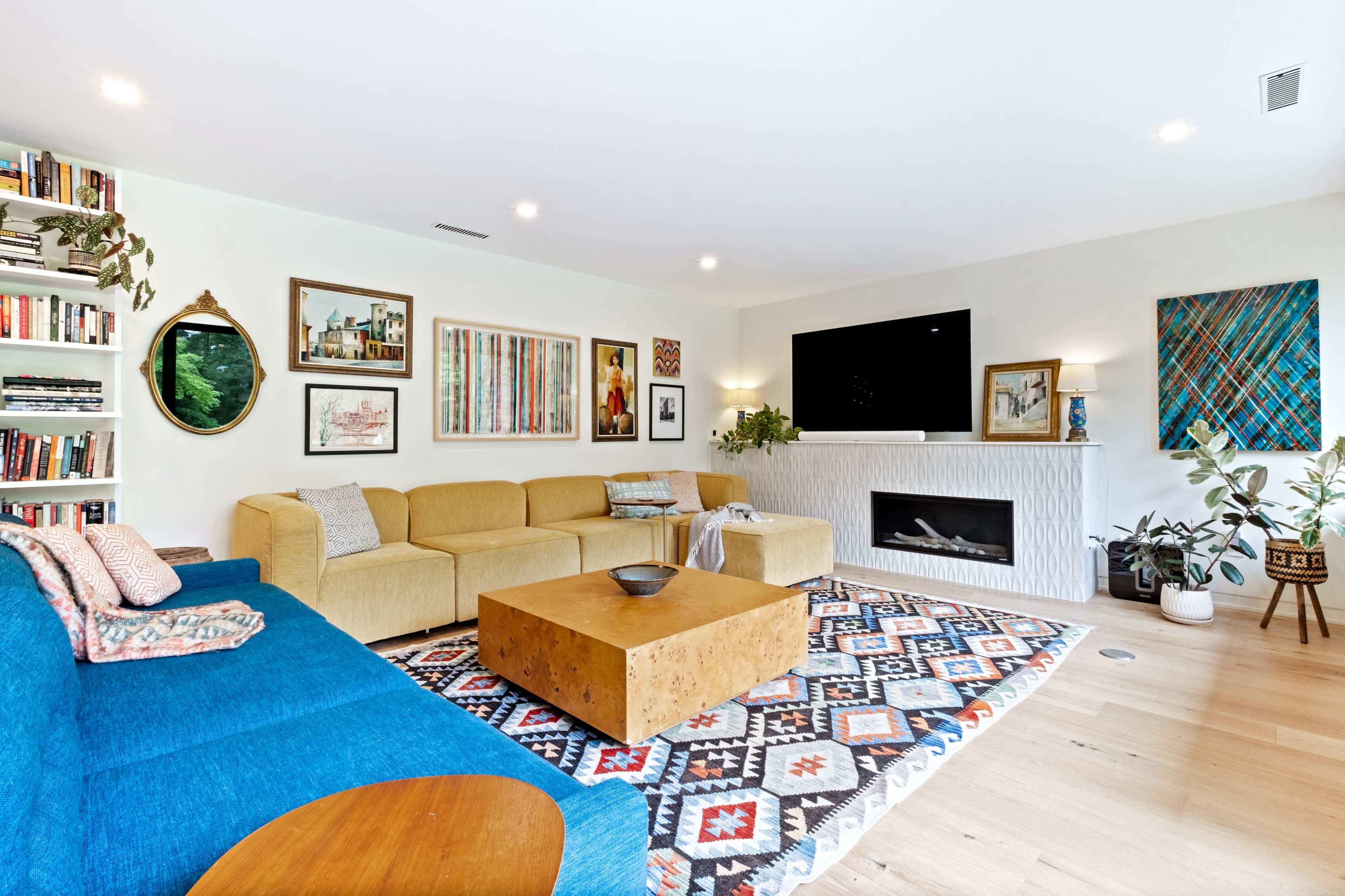 A modern living room features a blue couch, a yellow sectional, a wooden coffee table, a patterned area rug, mounted artworks, and a television above a white fireplace.