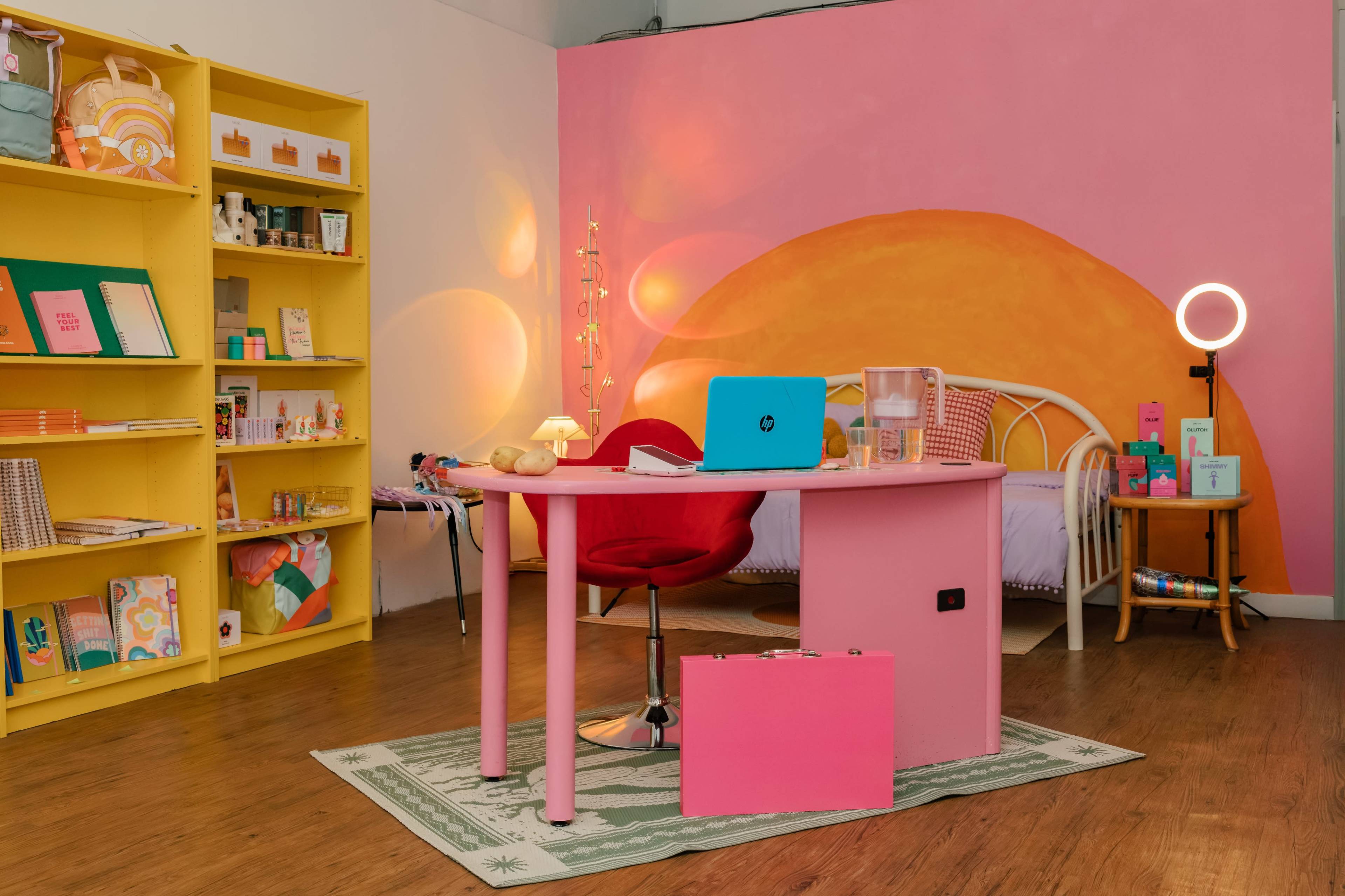 A brightly colored room features a pink desk and chair, a bed with a yellow wall mural, and shelves filled with various products.