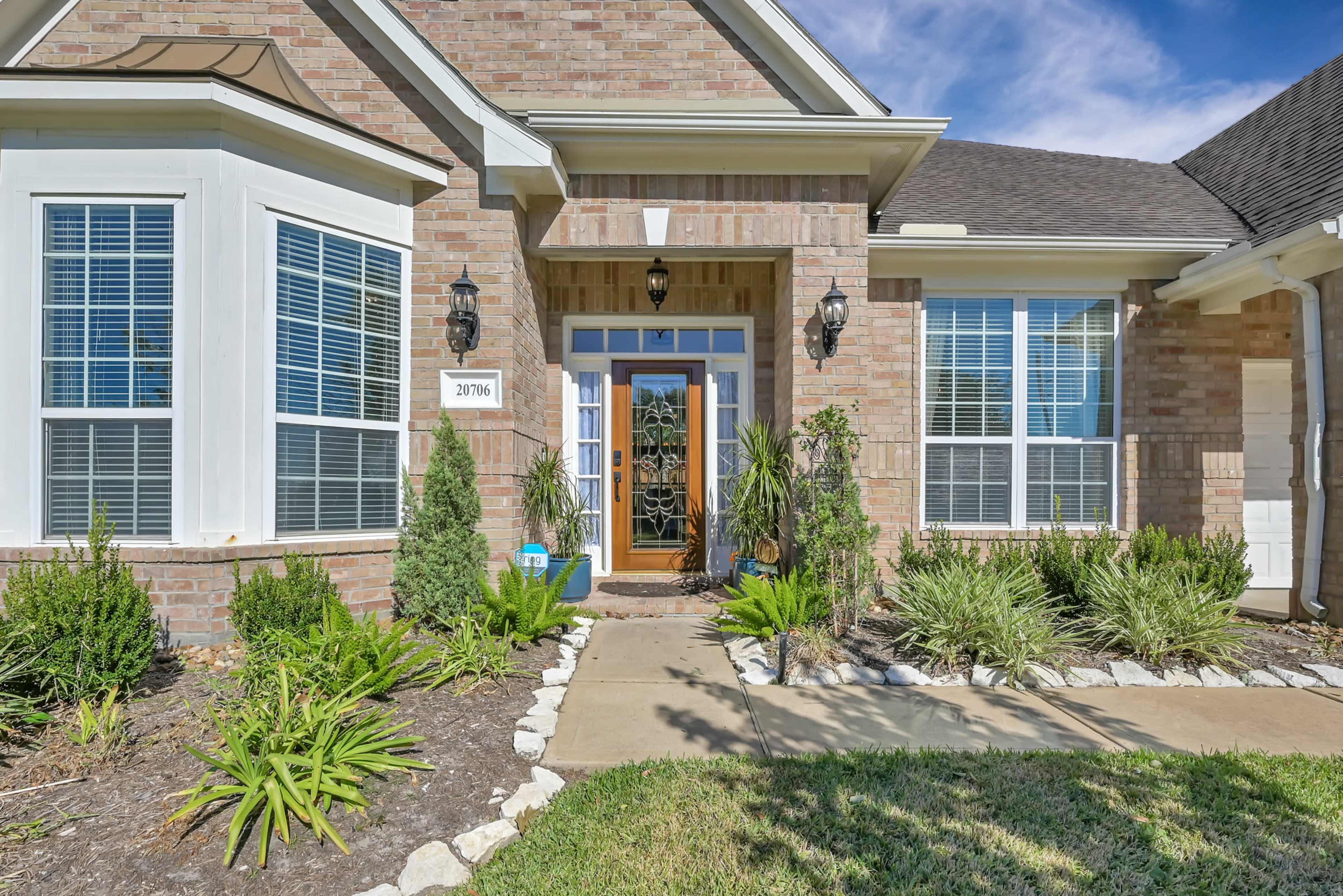 The entrance of a brick house with large windows, a wooden door, and landscaped gardens featuring various plants and shrubs.
