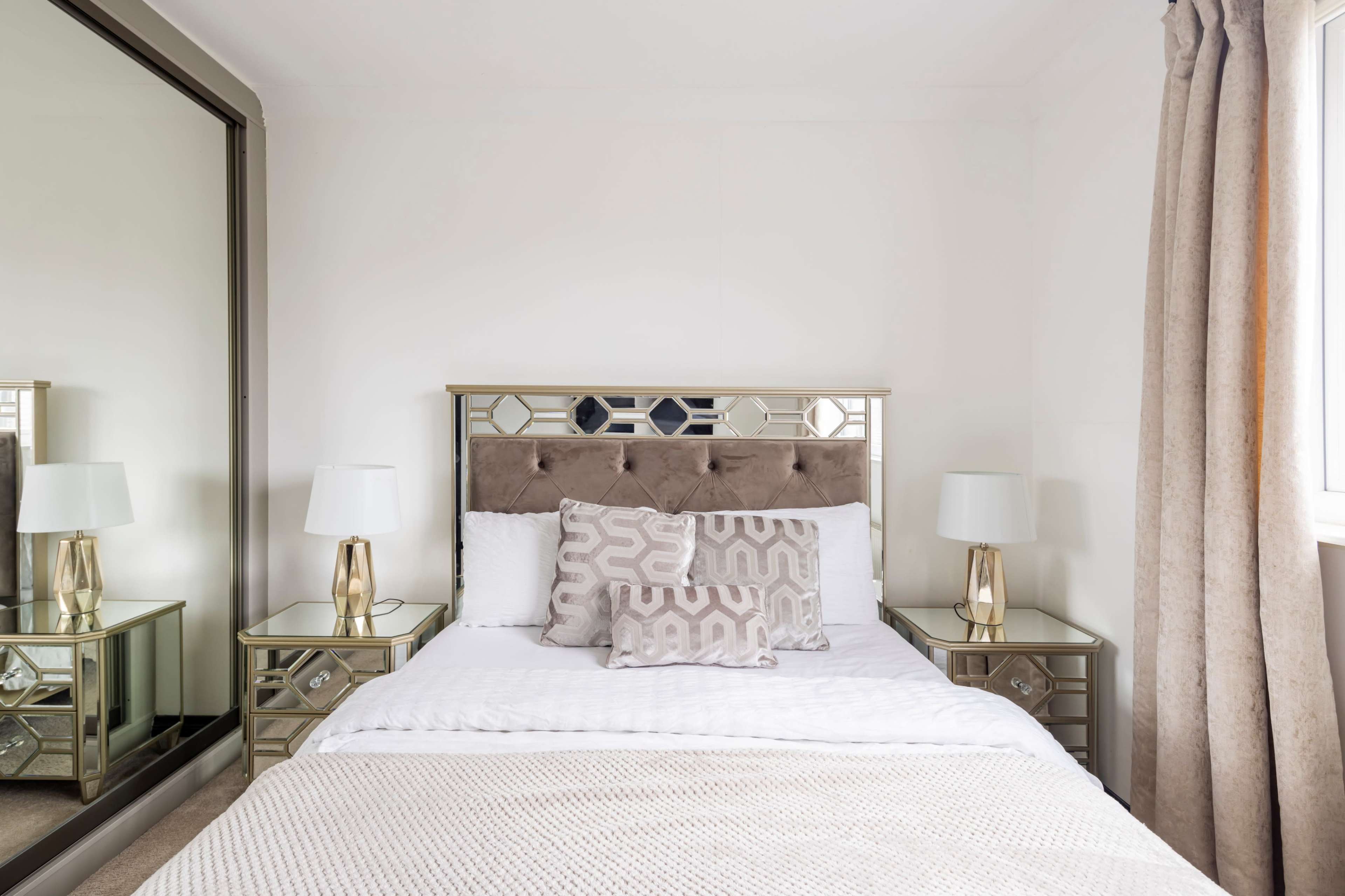 A neatly made bed with a tufted headboard and decorative pillows is positioned against a white wall, flanked by mirrored nightstands and lamps.