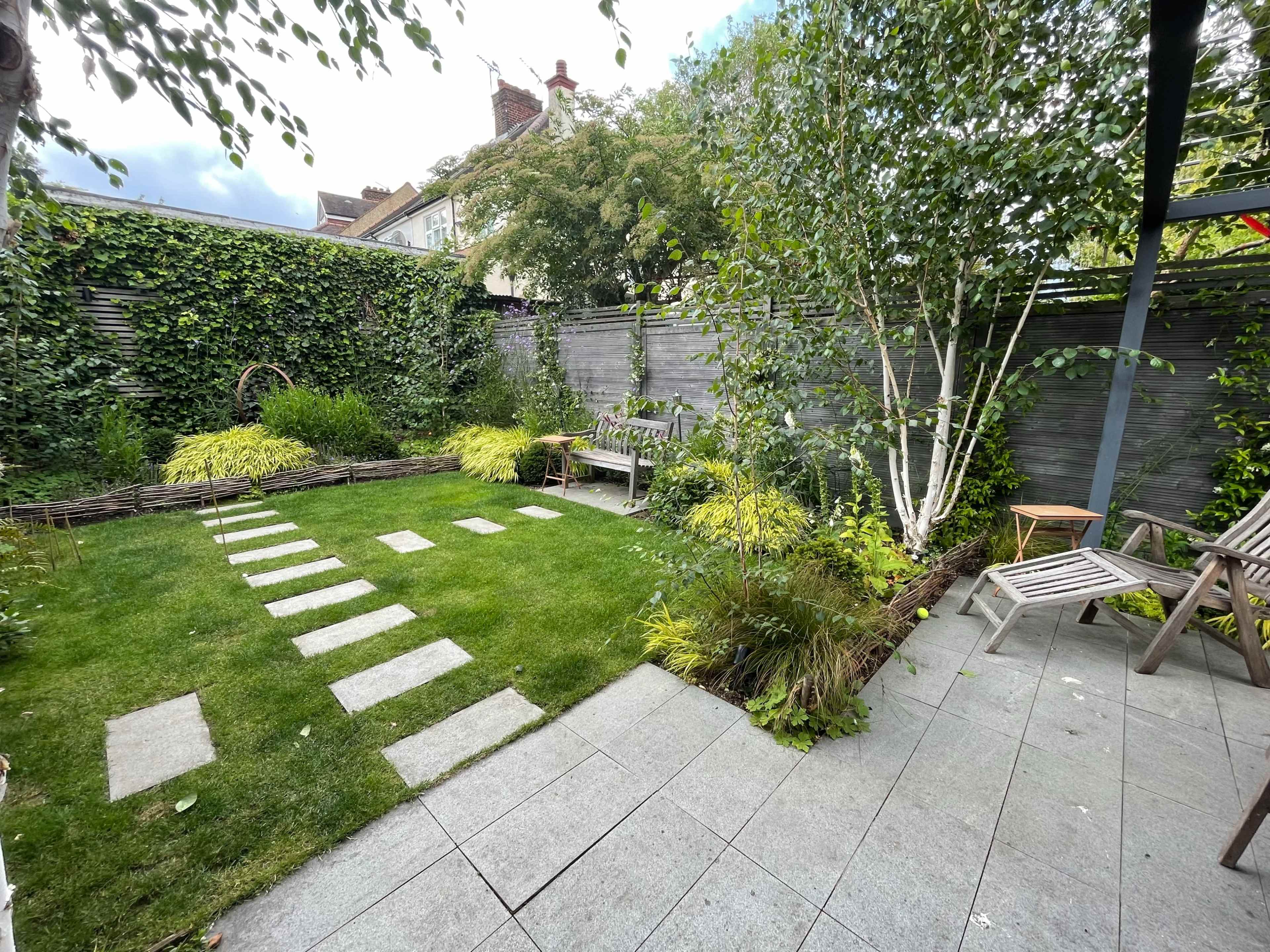 A landscaped garden featuring a rectangular lawn bordered by stone pathways and surrounded by various plants and trees.