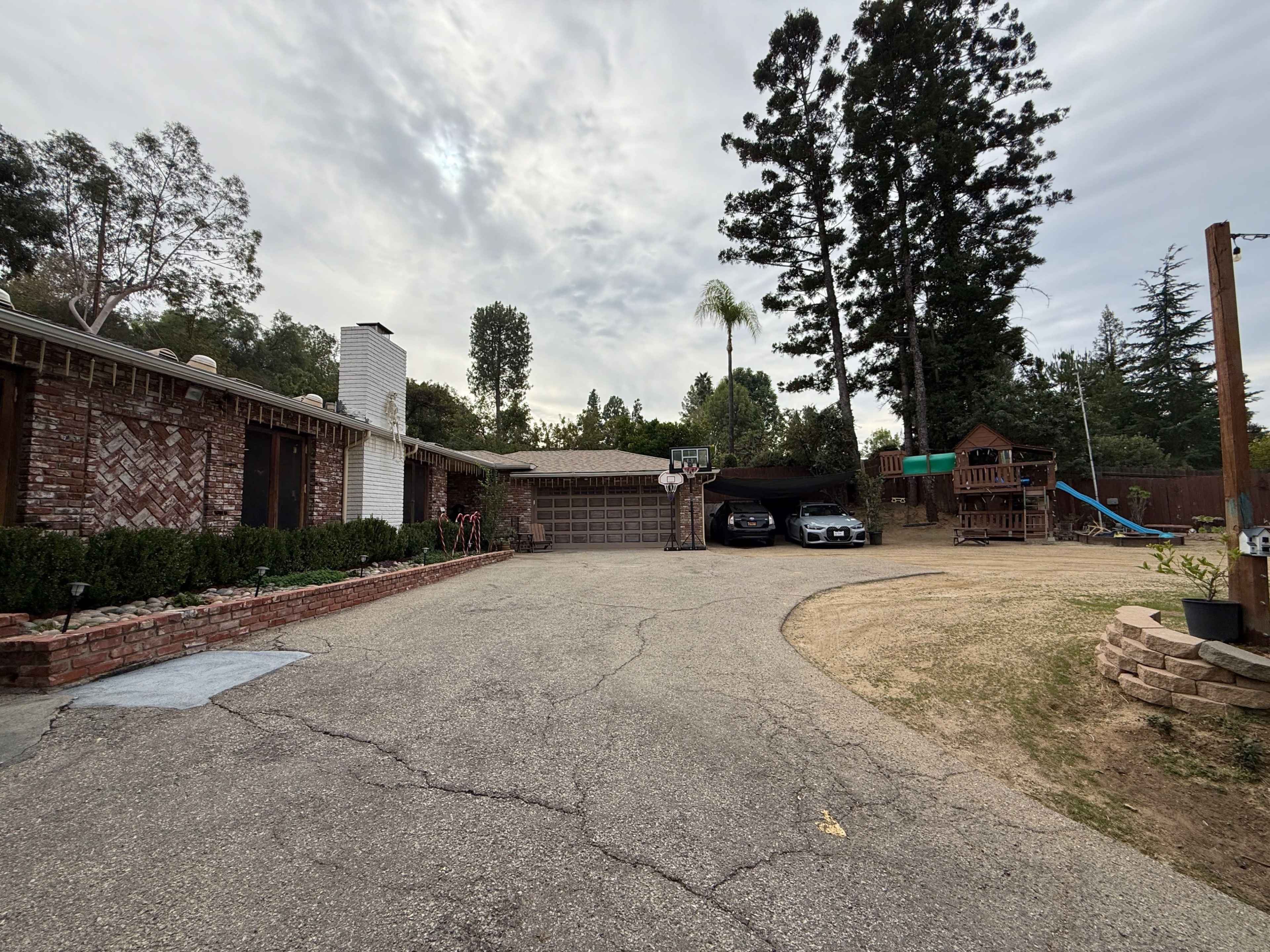 The image shows a residential driveway with a brick house on the left, a parked car, a basketball hoop, and playground equipment in a backyard.