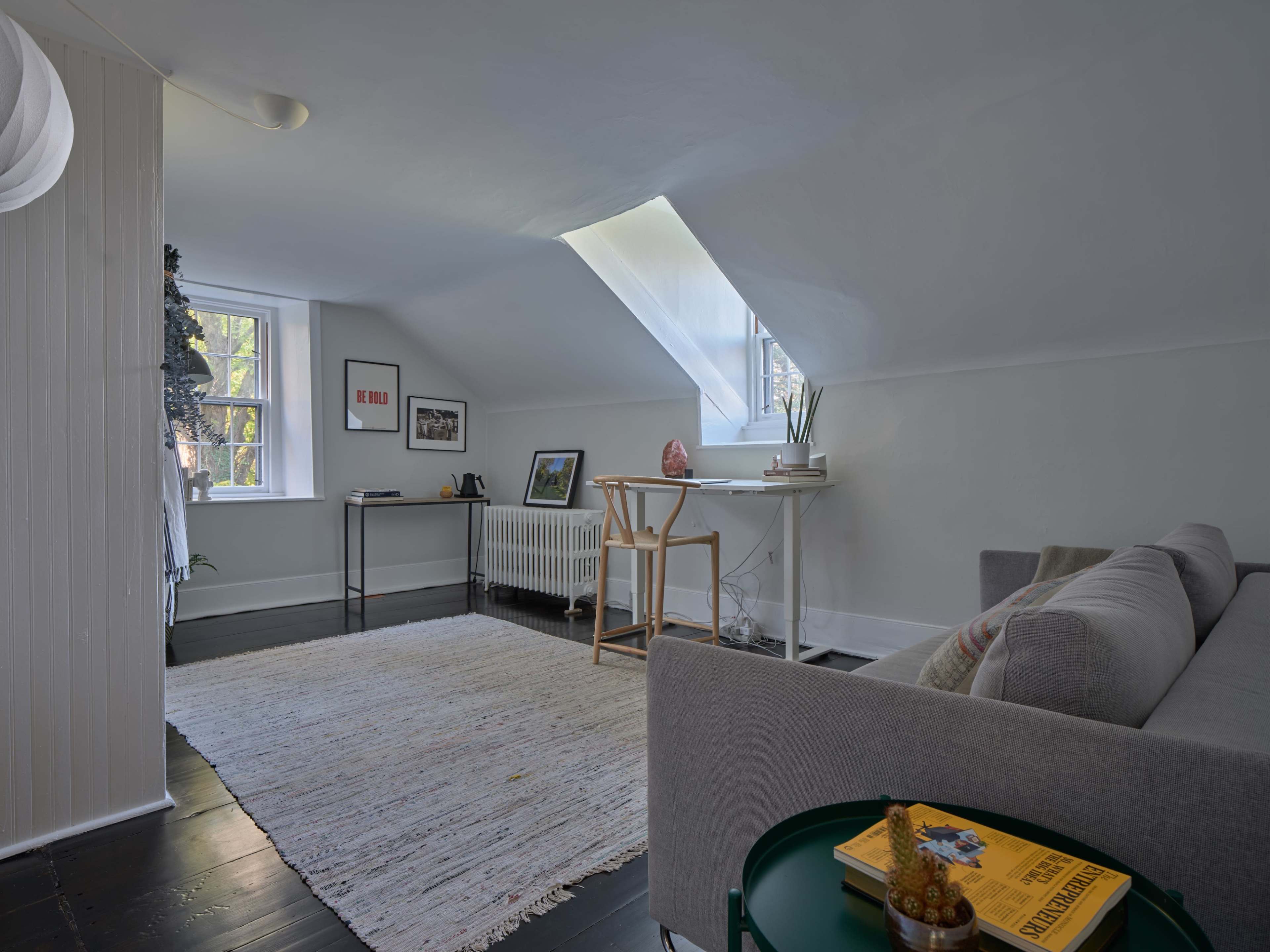 The room features a gray sofa, a small table, and a workspace under a skylight, with light-colored walls and a large area rug.