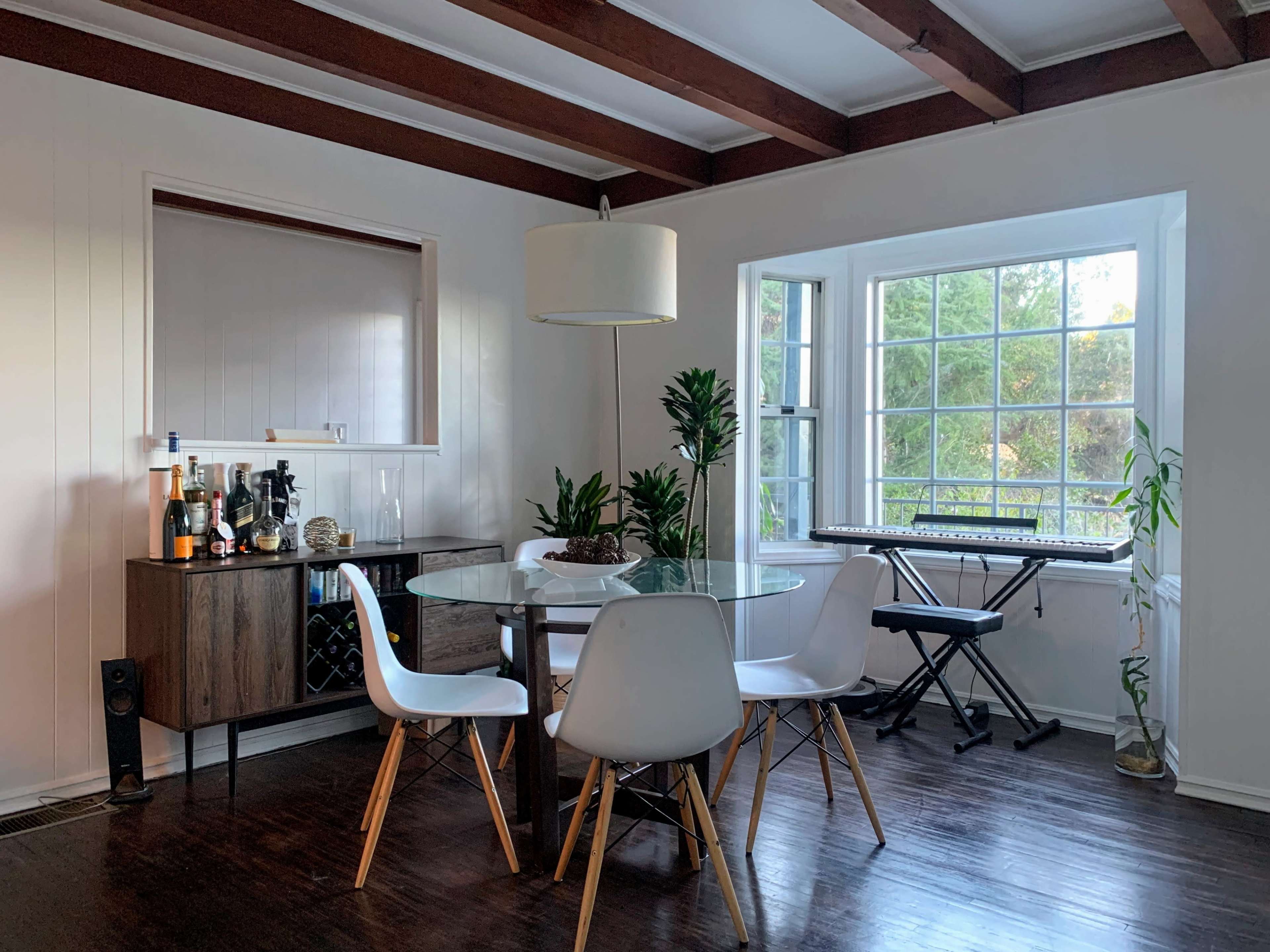 A dining area features a round glass table surrounded by white chairs, with a sideboard displaying drinks and a keyboard positioned near a large window.