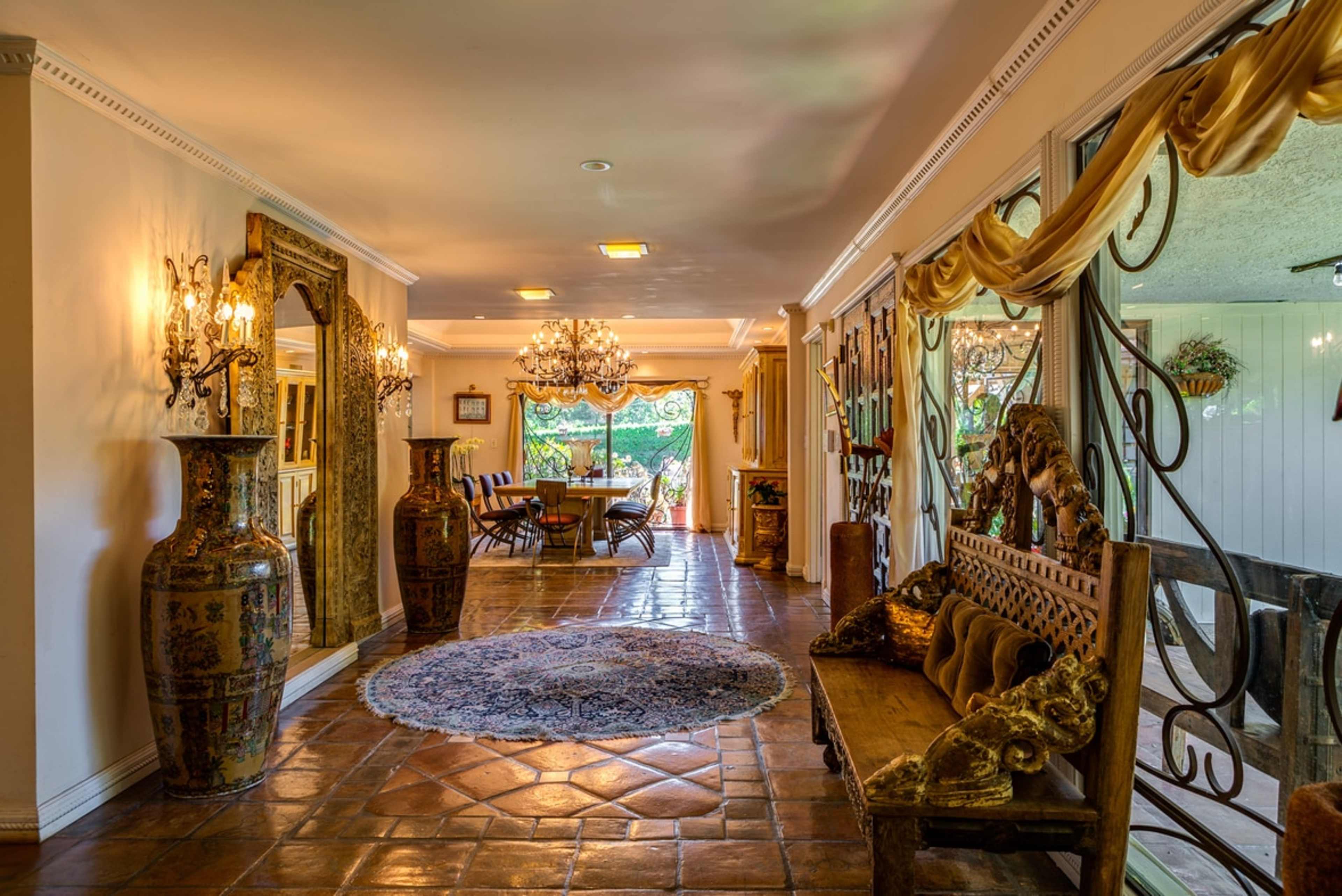 A spacious living area with a decorative rug, two large vases, a wooden bench, and a dining table set under elegant chandeliers.