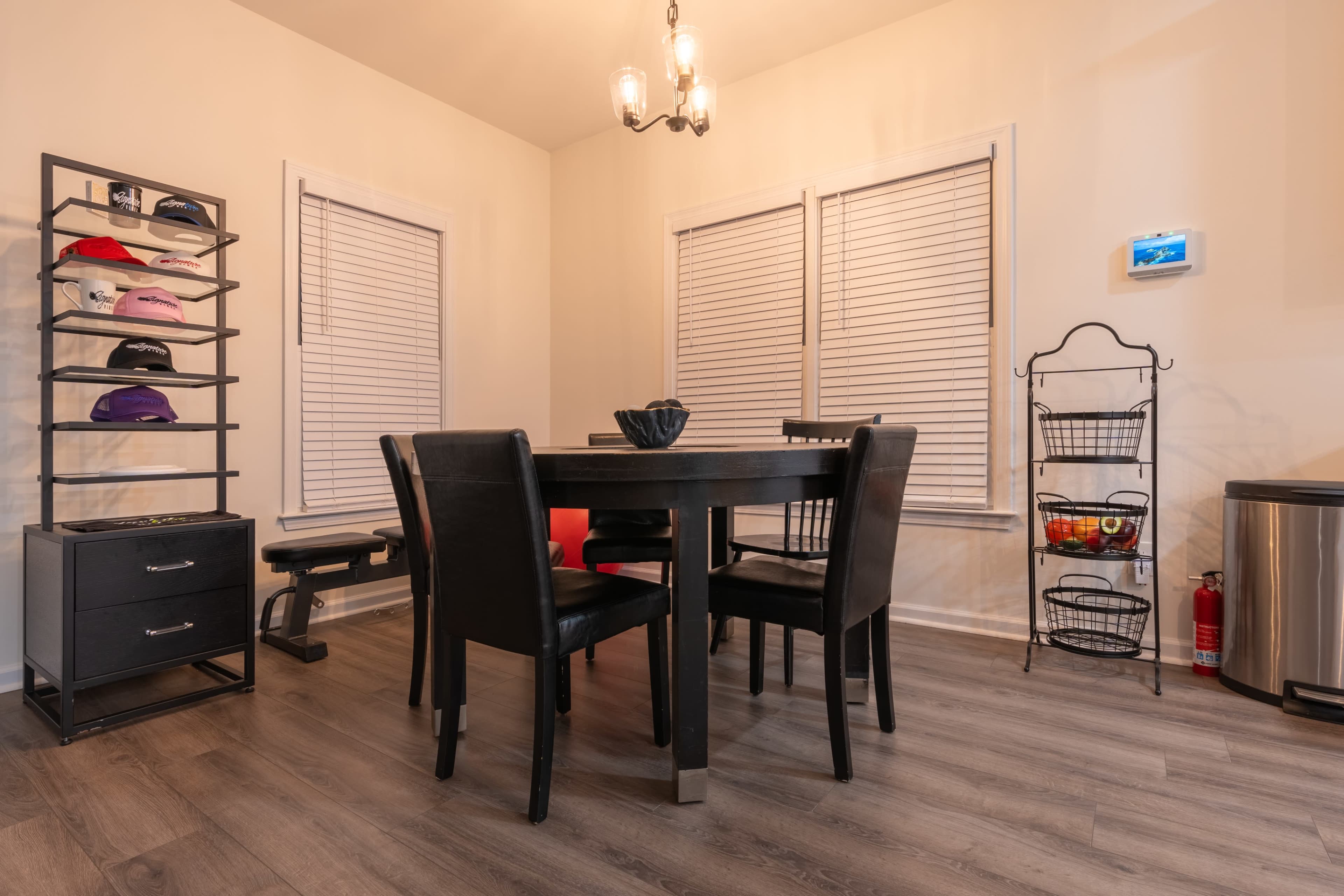A dining area features a black table with four chairs, a shelving unit displaying caps, and a storage cart next to a window with blinds.
