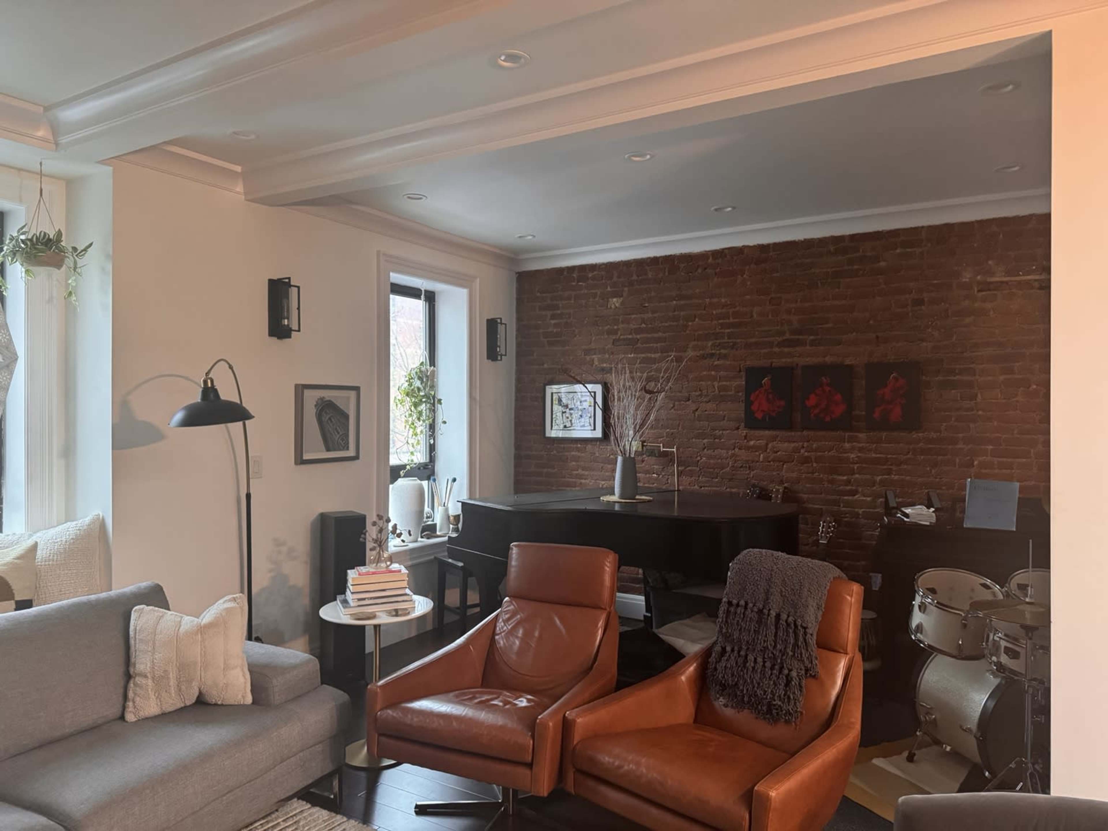 The image shows a cozy living space with a gray sofa, two brown leather chairs, a piano against a brick wall, and a small drum set in the corner.