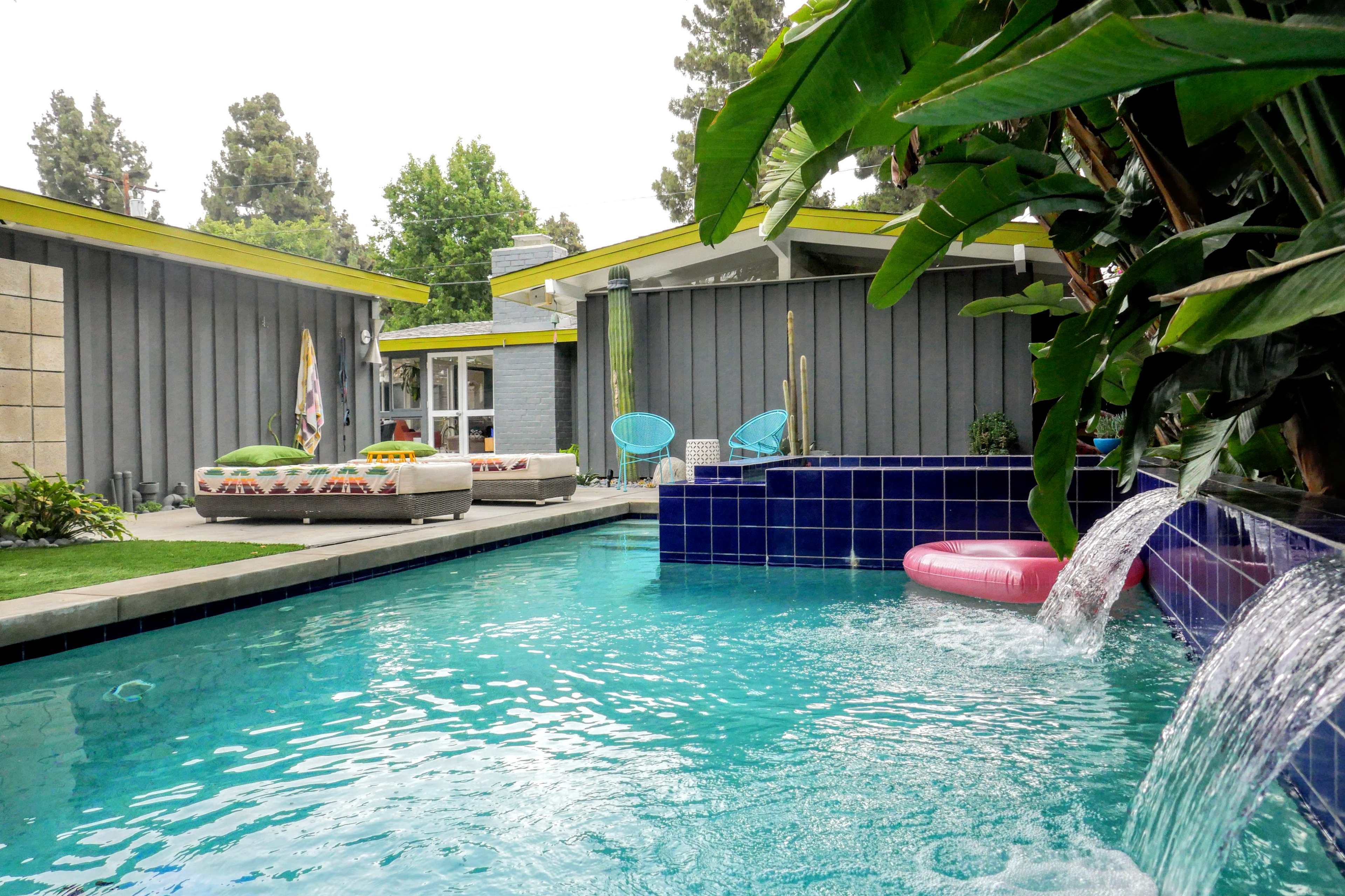 The image shows a backyard with a swimming pool, a waterfall feature, lounge furniture, and palm trees against a modern home facade.