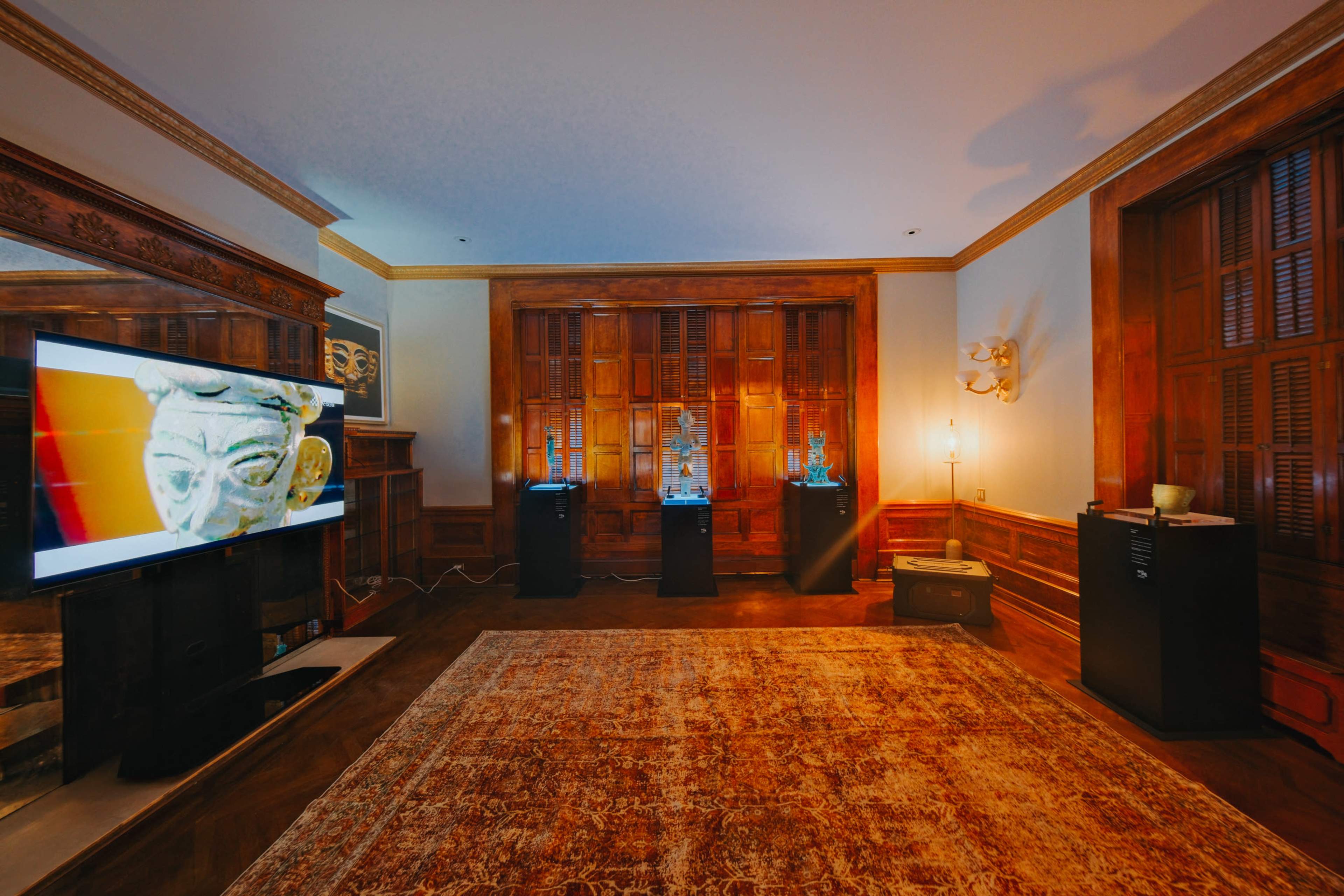 The scene features a wooden room with a patterned rug, a television displaying an image, and three display stands showcasing artifacts against a backdrop of wooden paneling.