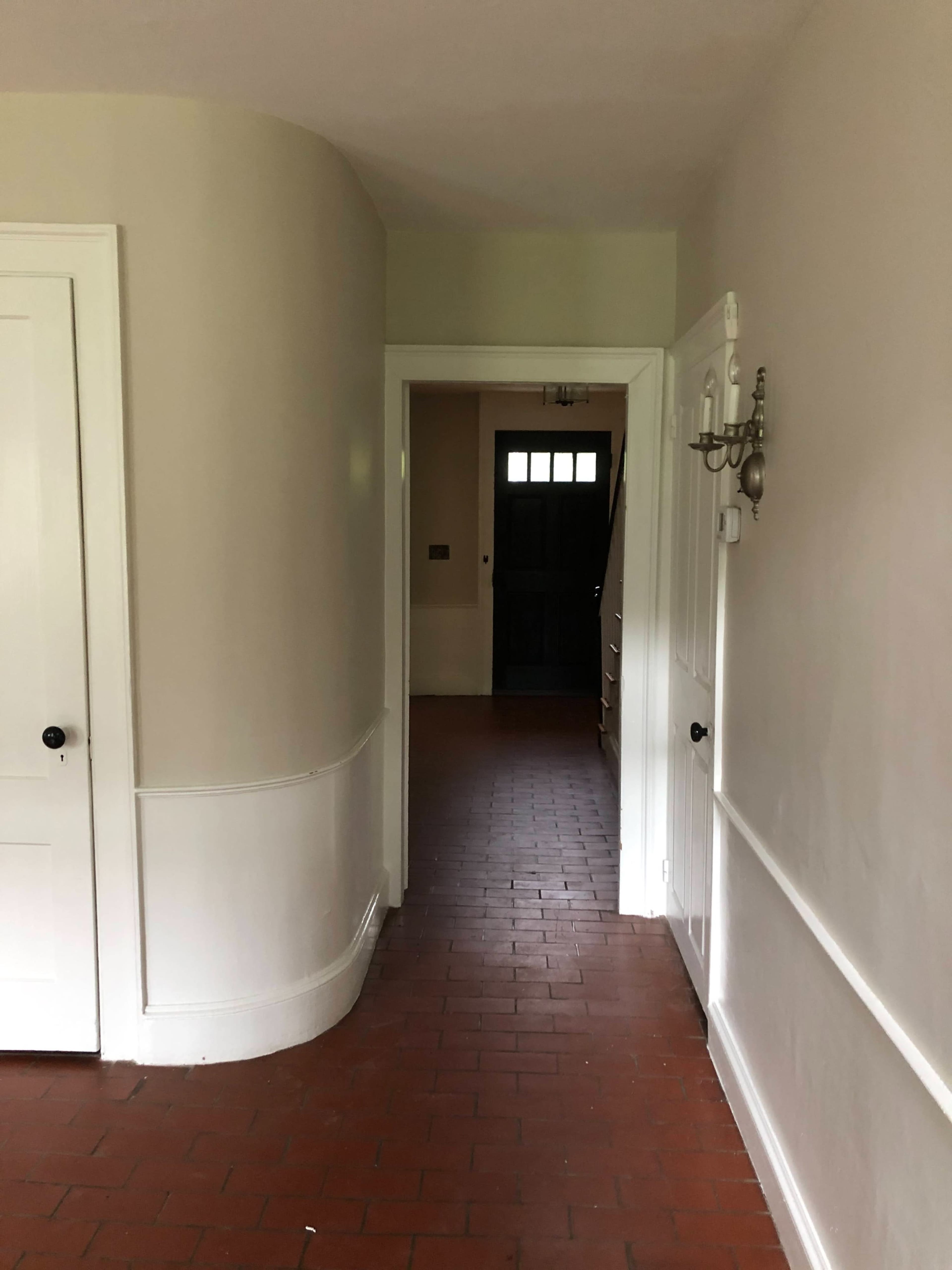 A narrow hallway with a curved wall leading to a door at the end, featuring brick flooring and white paneling.