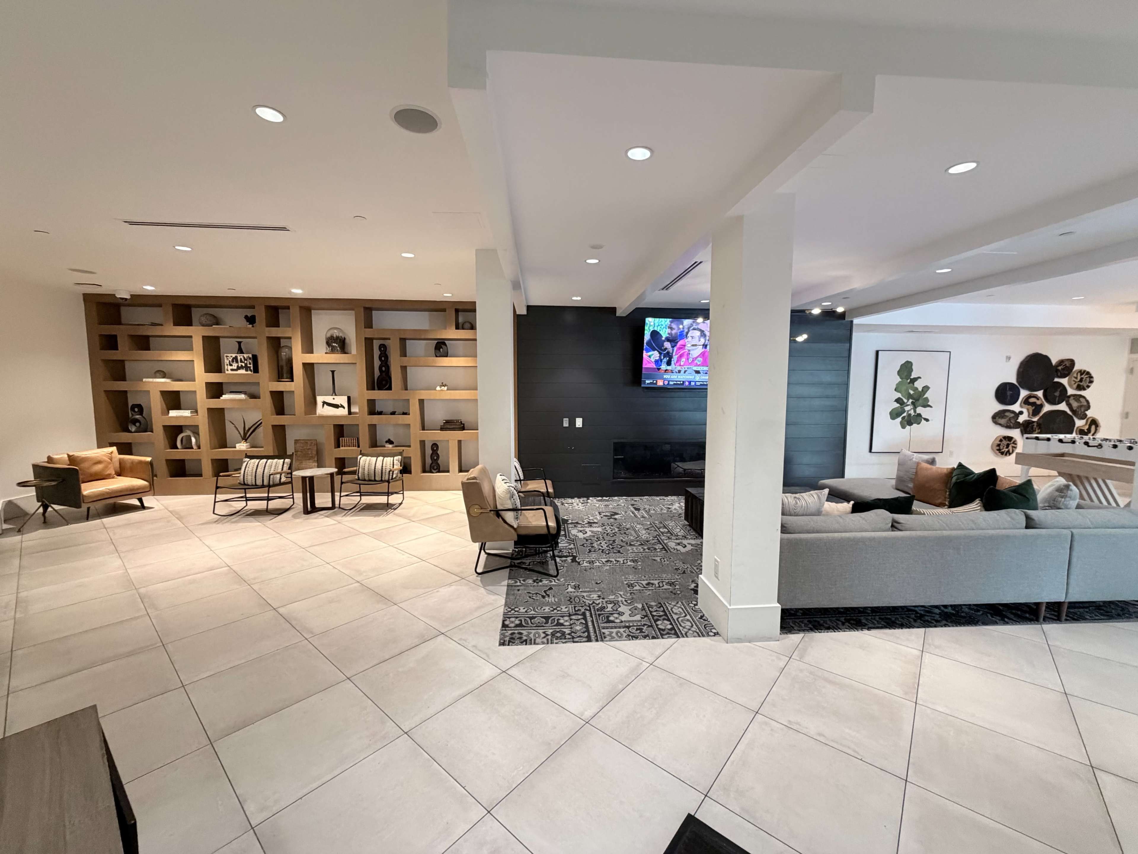 The image shows a modern lounge area with a variety of seating options, a television on the wall, and decorative shelving.