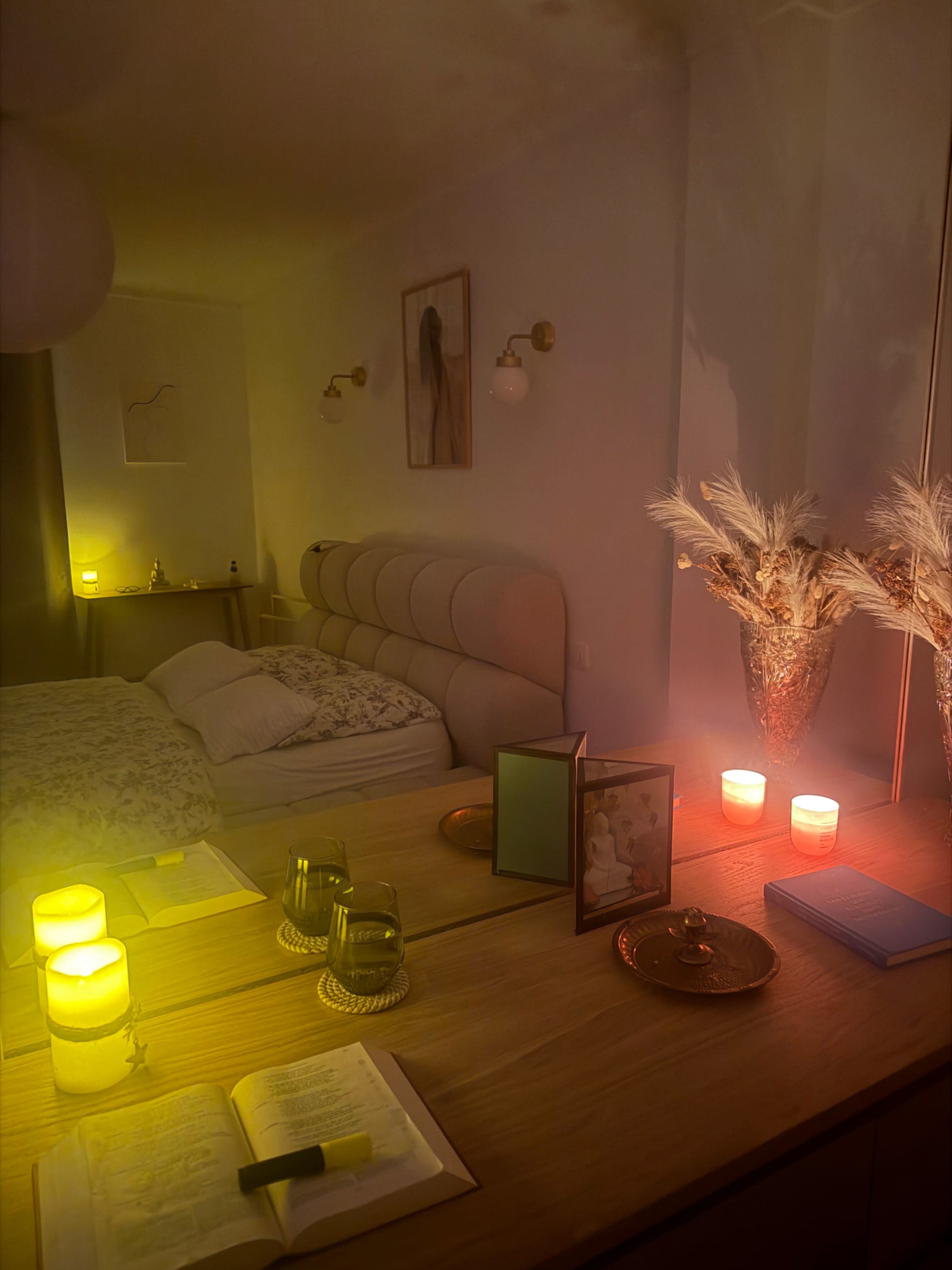A softly lit bedroom features a neatly made bed, a dresser with candles and decorative items, and a reflection in the mirror that highlights the warm glow of the ambient lighting.