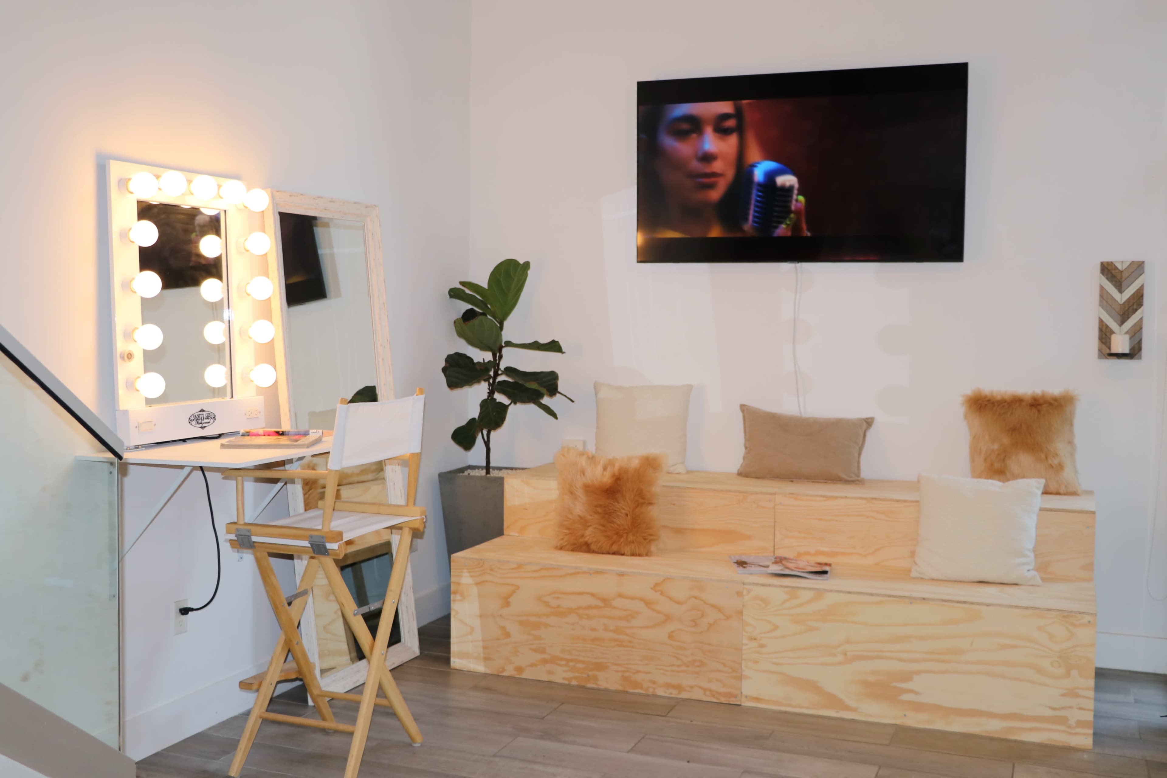 The scene features a stylish vanity area with a mirror surrounded by lights, a television on the wall, and a seating space made of wooden platforms with decorative pillows.