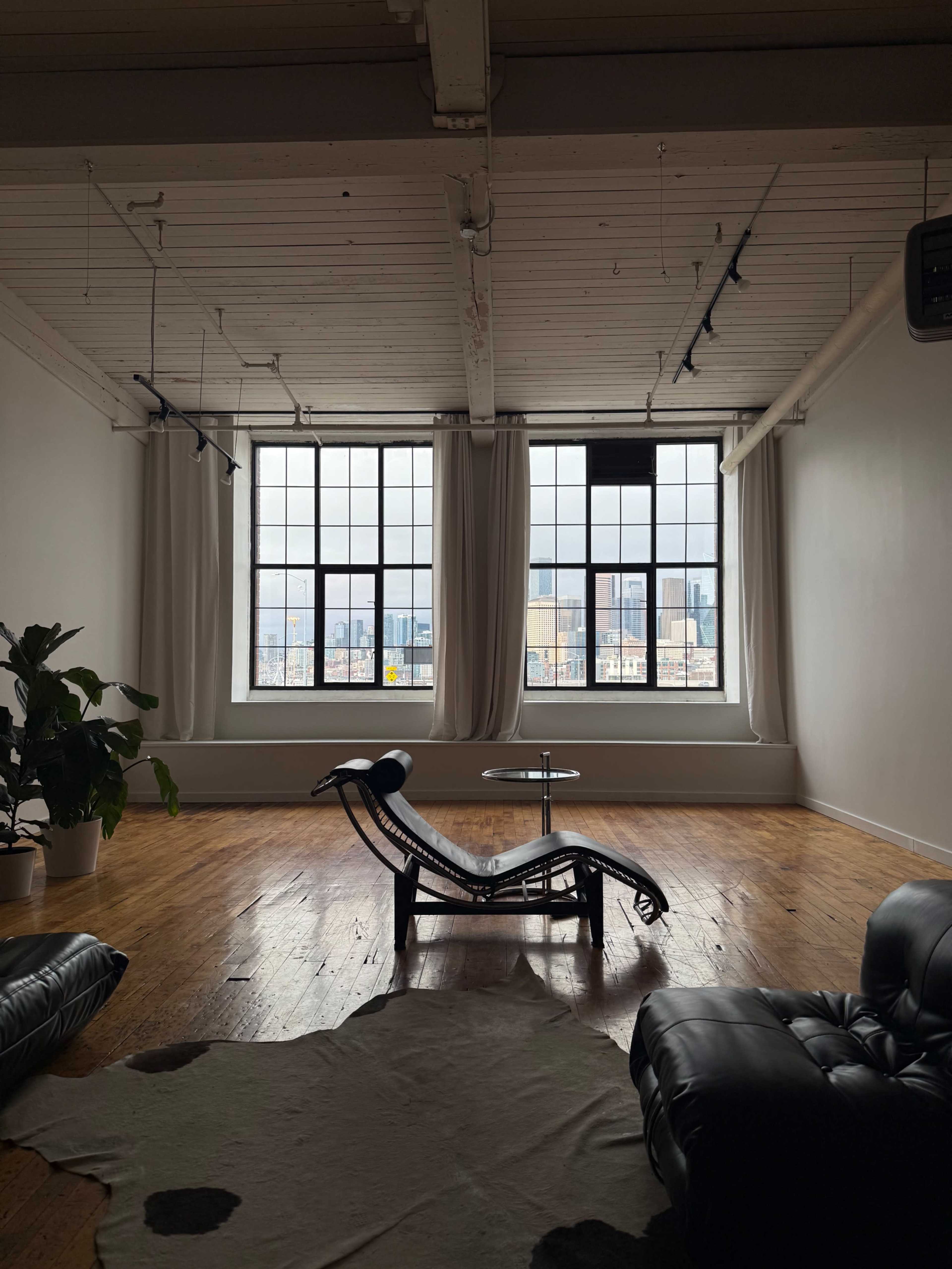The image shows a spacious room with large windows, a chaise lounge, a small table, and a potted plant on the floor.