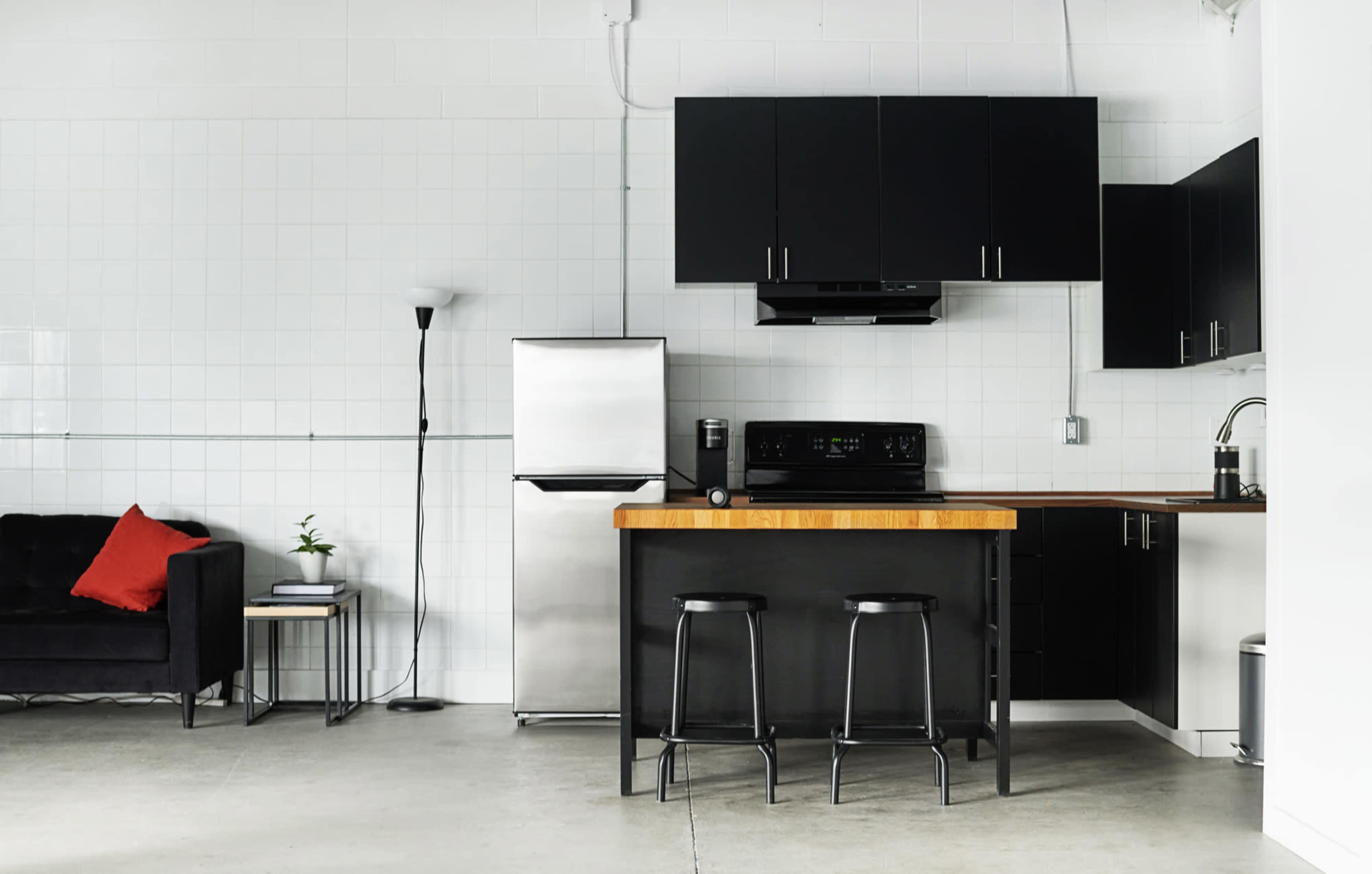 A modern kitchen with a stainless steel refrigerator, black cabinetry, a wooden countertop with two stools, and a black sofa with a red pillow.