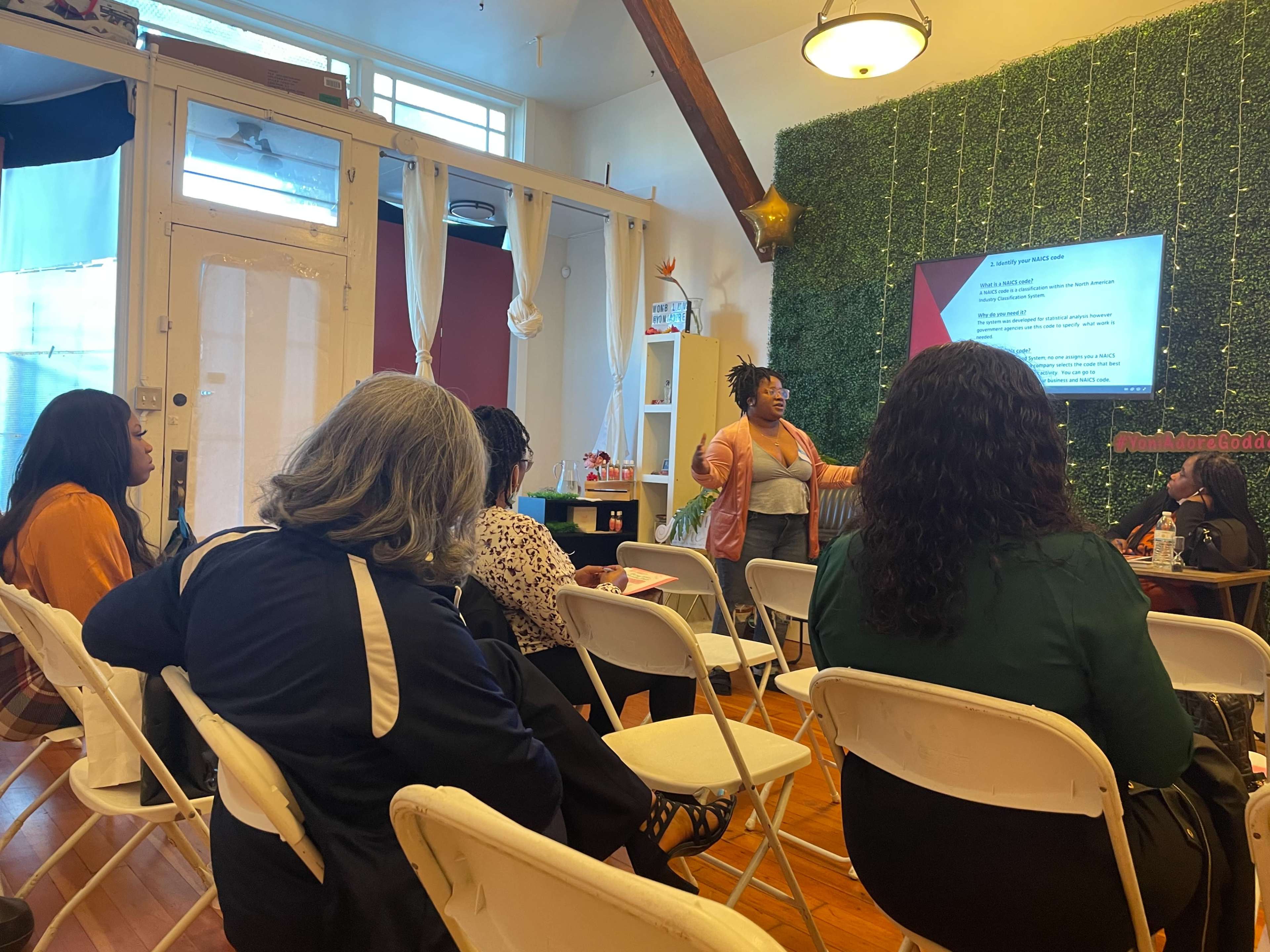 A group of people sit in folding chairs facing a speaker during a presentation in a brightly lit room with green decor on the walls.