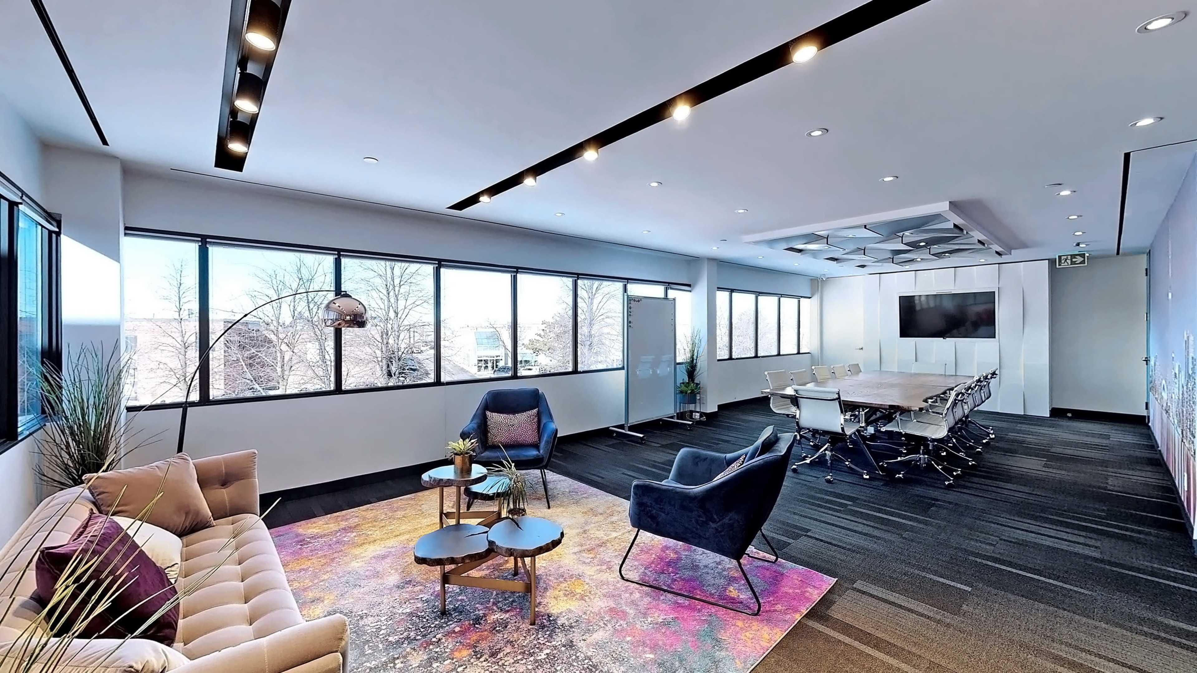 The image depicts a modern conference room with large windows, showcasing a seating area with a sofa and armchairs, a coffee table, and a long meeting table surrounded by ergonomic chairs.