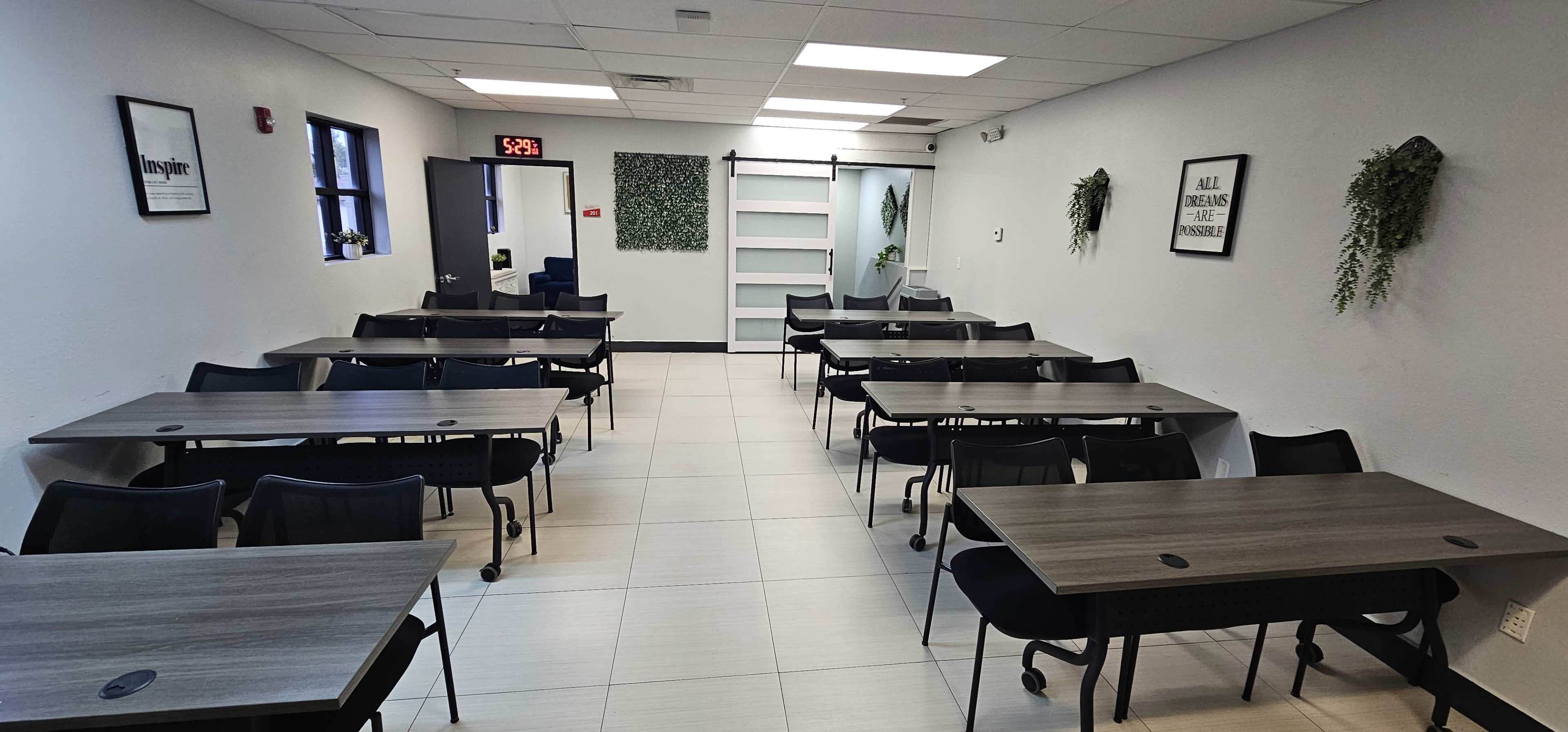 A brightly lit classroom features several rows of gray tables and black chairs arranged for seating, with a wall of greenery and shelves at one end.