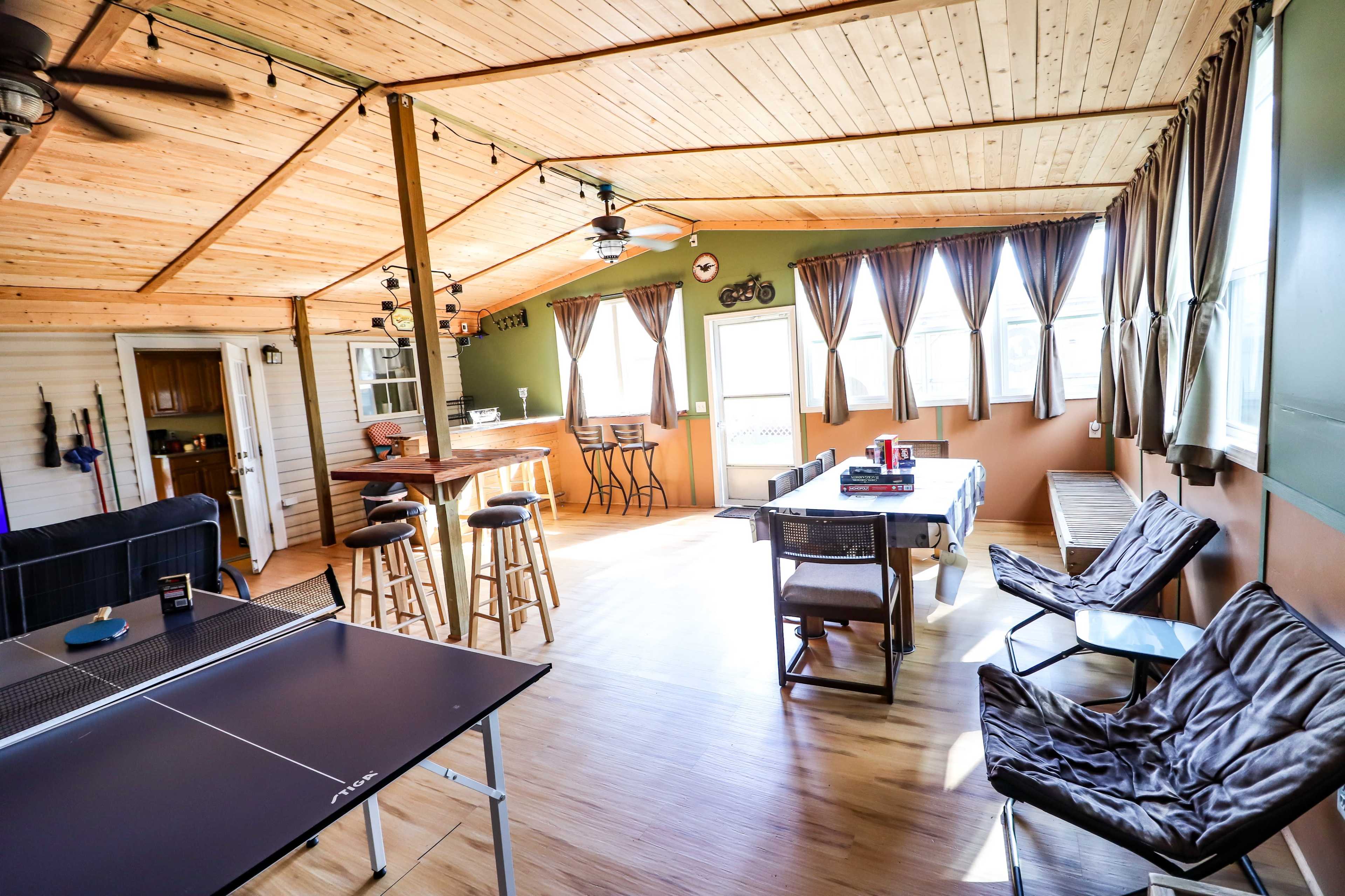 The image showcases a spacious room with wooden ceilings, a dining table, bar stools, and a ping pong table, featuring large windows that allow natural light to enter.
