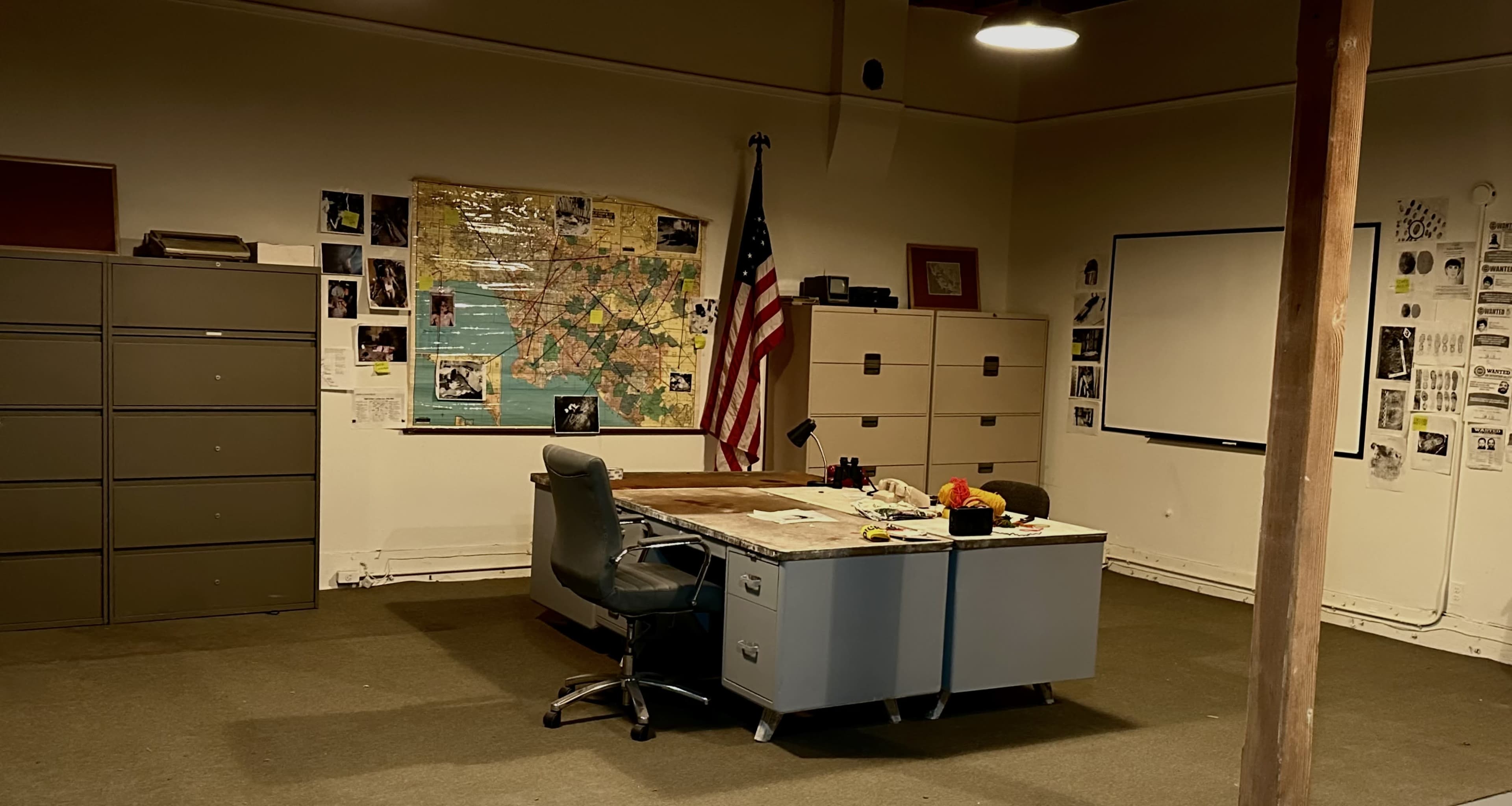 A sparsely furnished office with a large desk, an American flag, a map on the wall, and several filing cabinets.