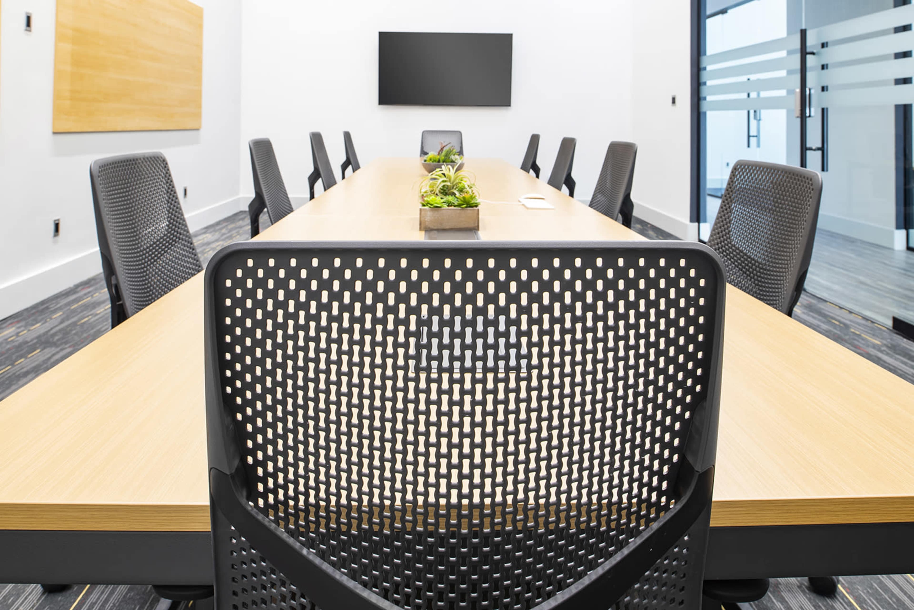 A long conference table with a row of black chairs and a small plant centerpiece is set up in a modern meeting room.