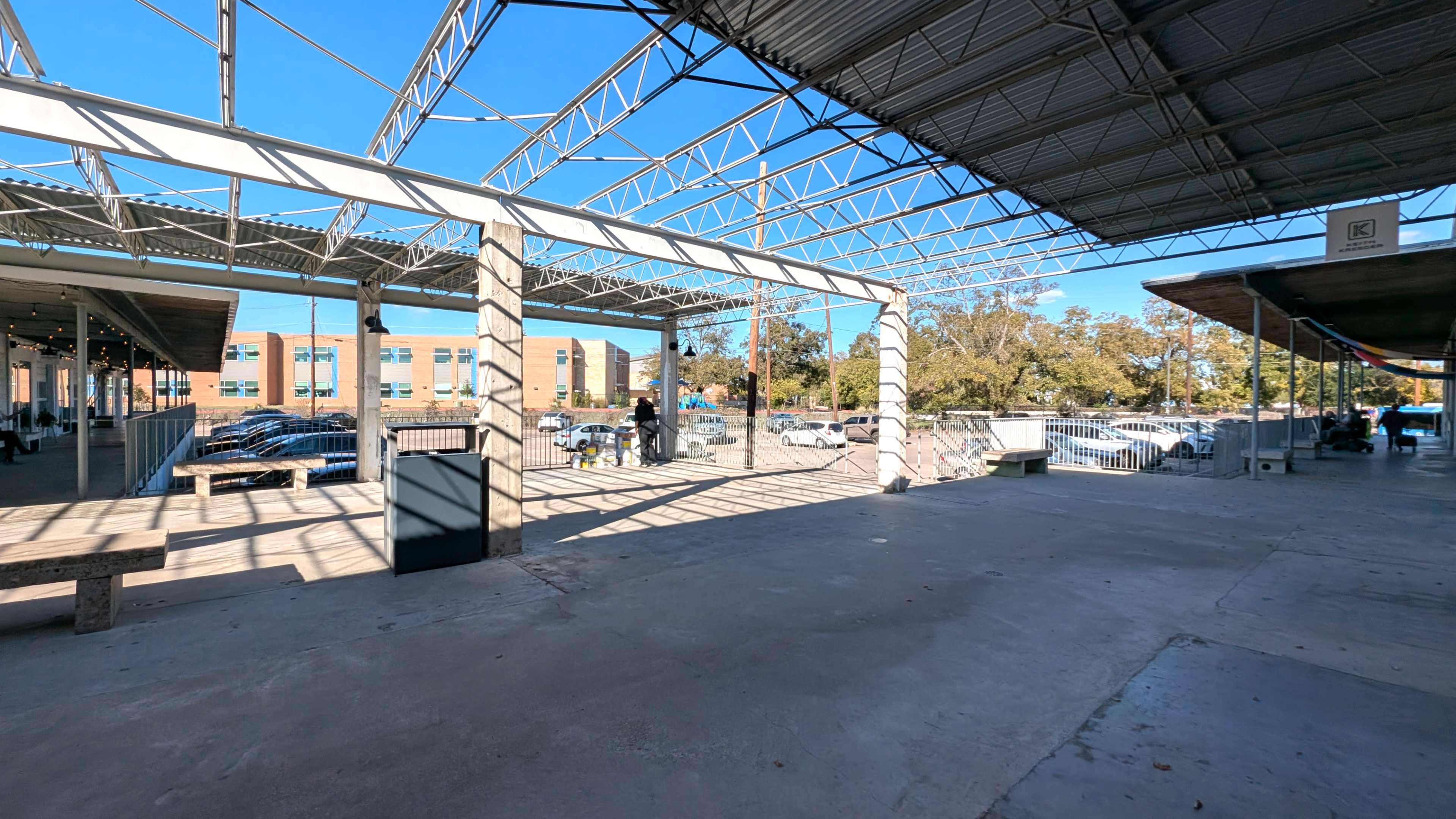 The image shows a spacious outdoor area with a metal roof structure, concrete floor, and several parked cars visible in the background.